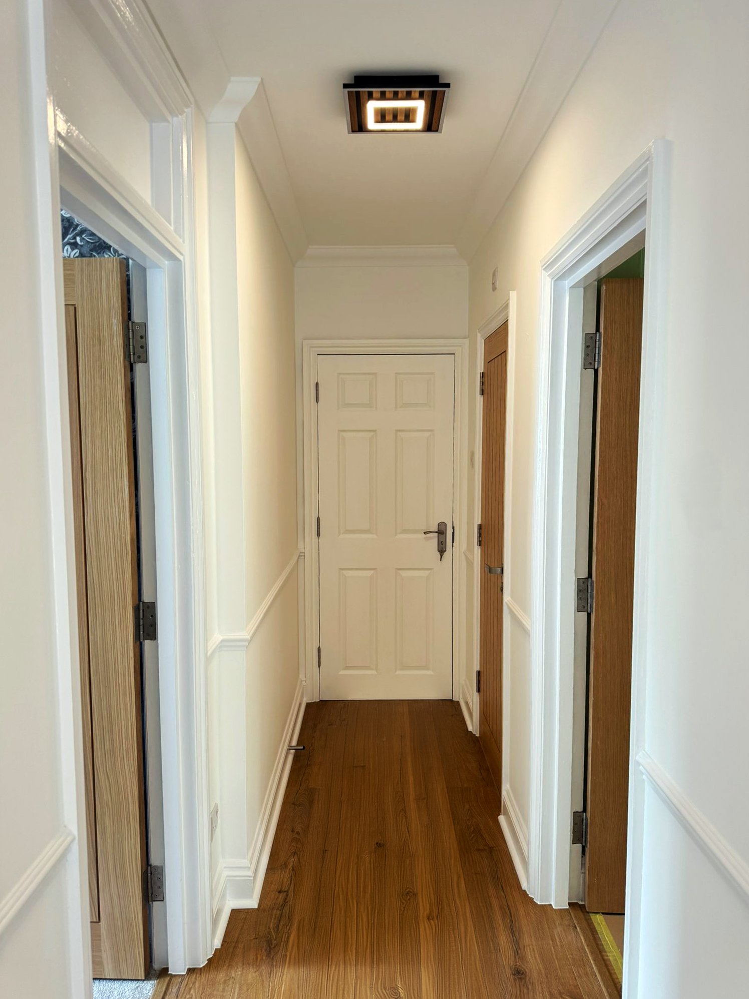 A hallway with white walls and trim, hardwood floor, and a modern square ceiling light fixture, with several doors along the corridor.