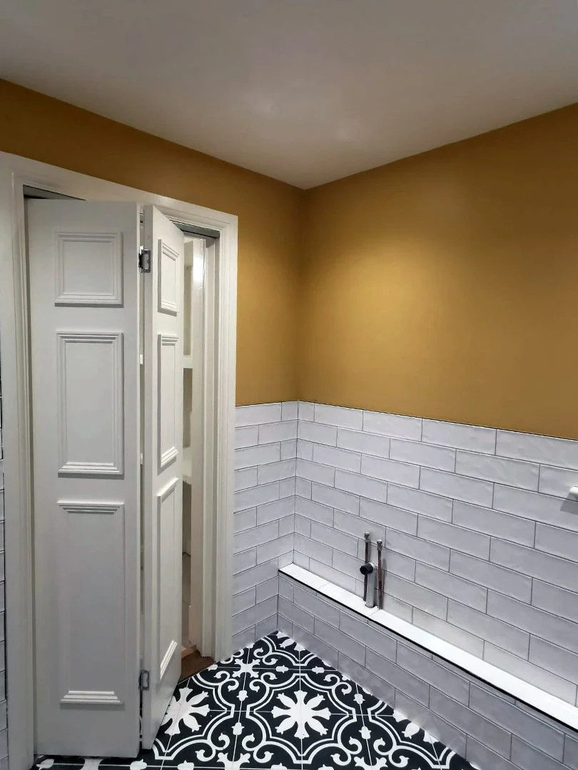 Bathroom with beige upper wall, white subway tile wainscoting, black and white patterned floor tile, and a white door with a folding cover.