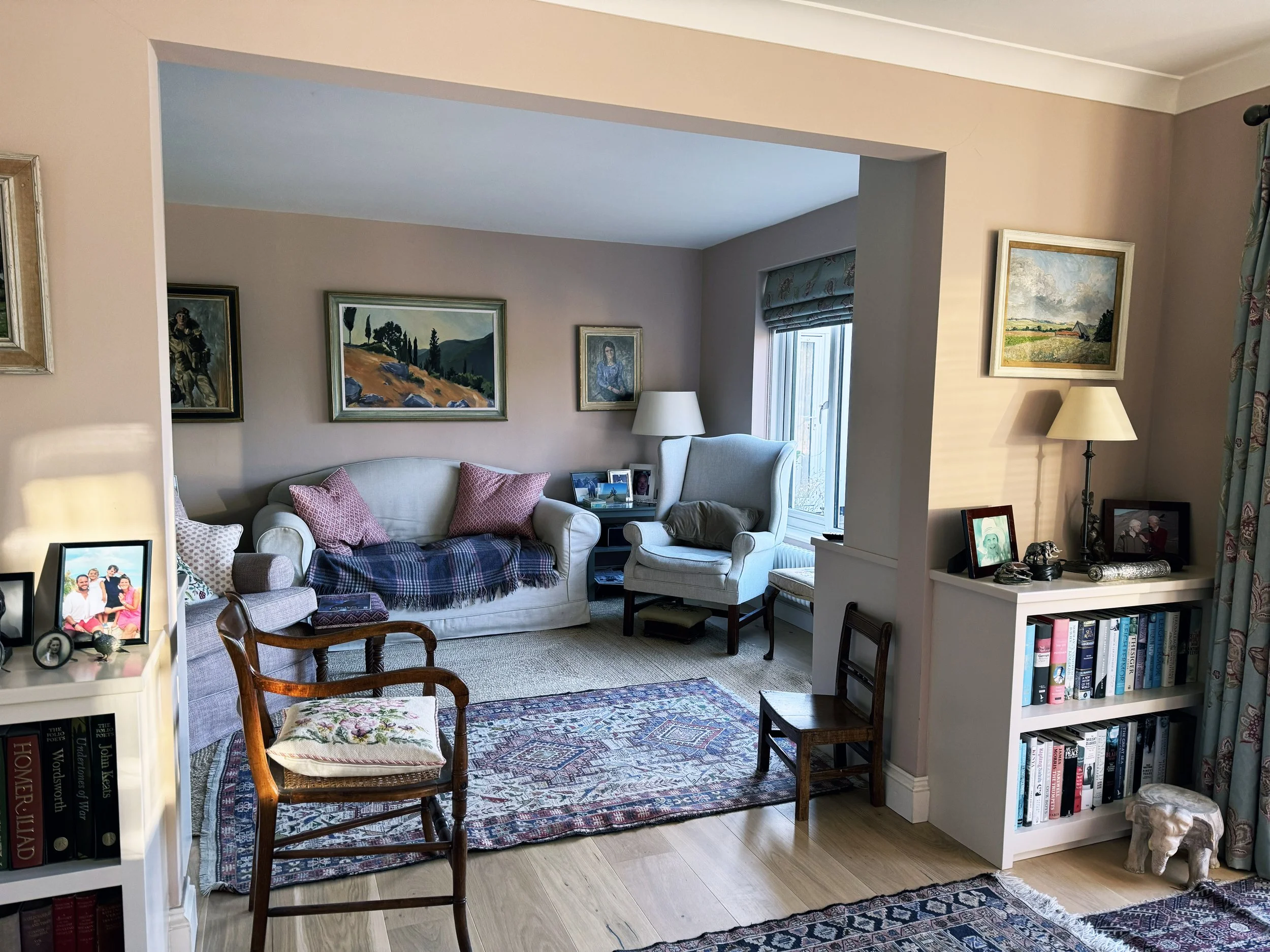 Living room with white couch, two armchairs, wooden chairs, a colorful area rug, bookshelves, paintings, and family photos.