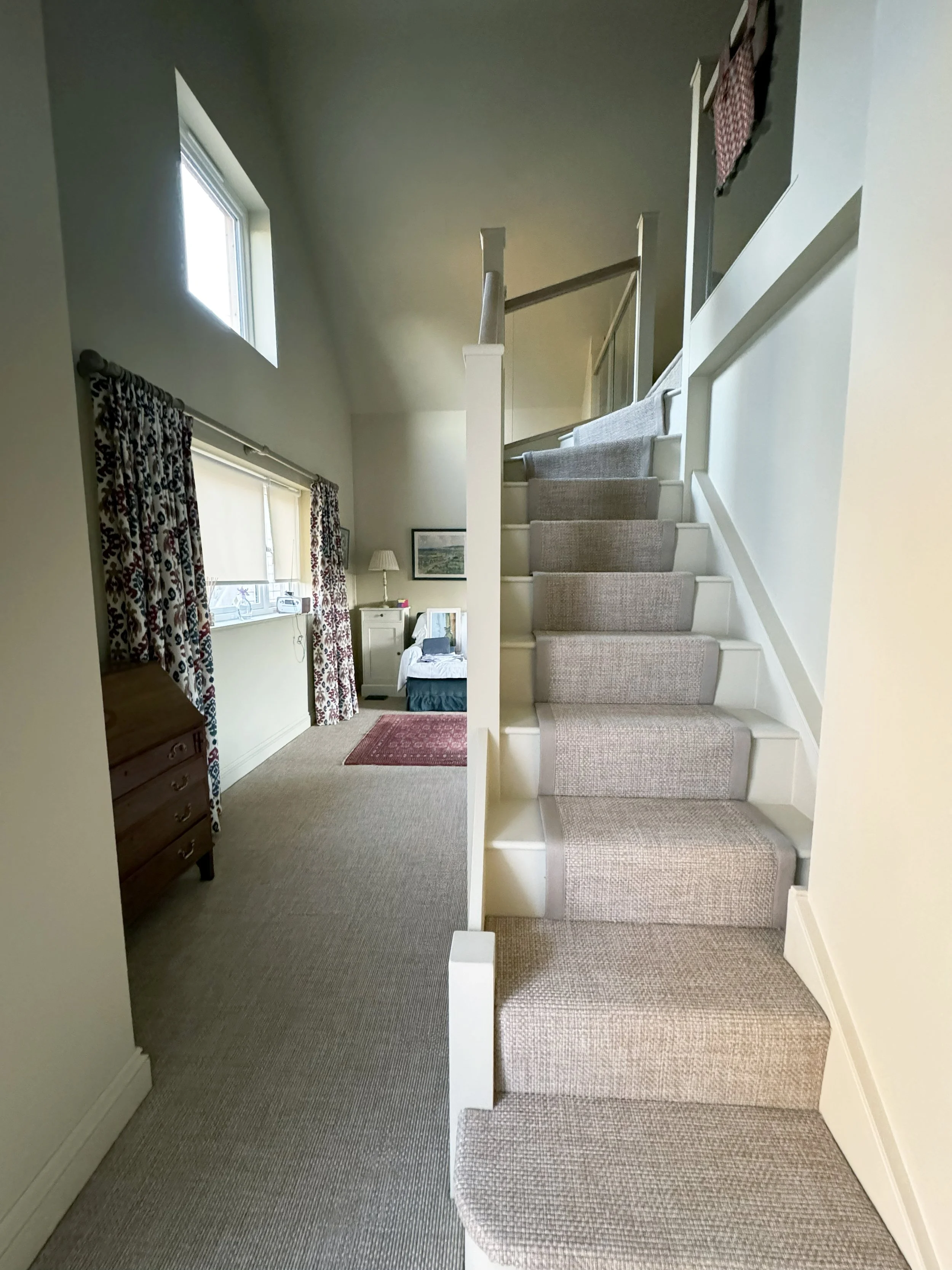 A staircase with beige carpet runner seen from below, leading upstairs in a home interior near a bedroom with patterned curtains, window, dresser, and wall decorations.