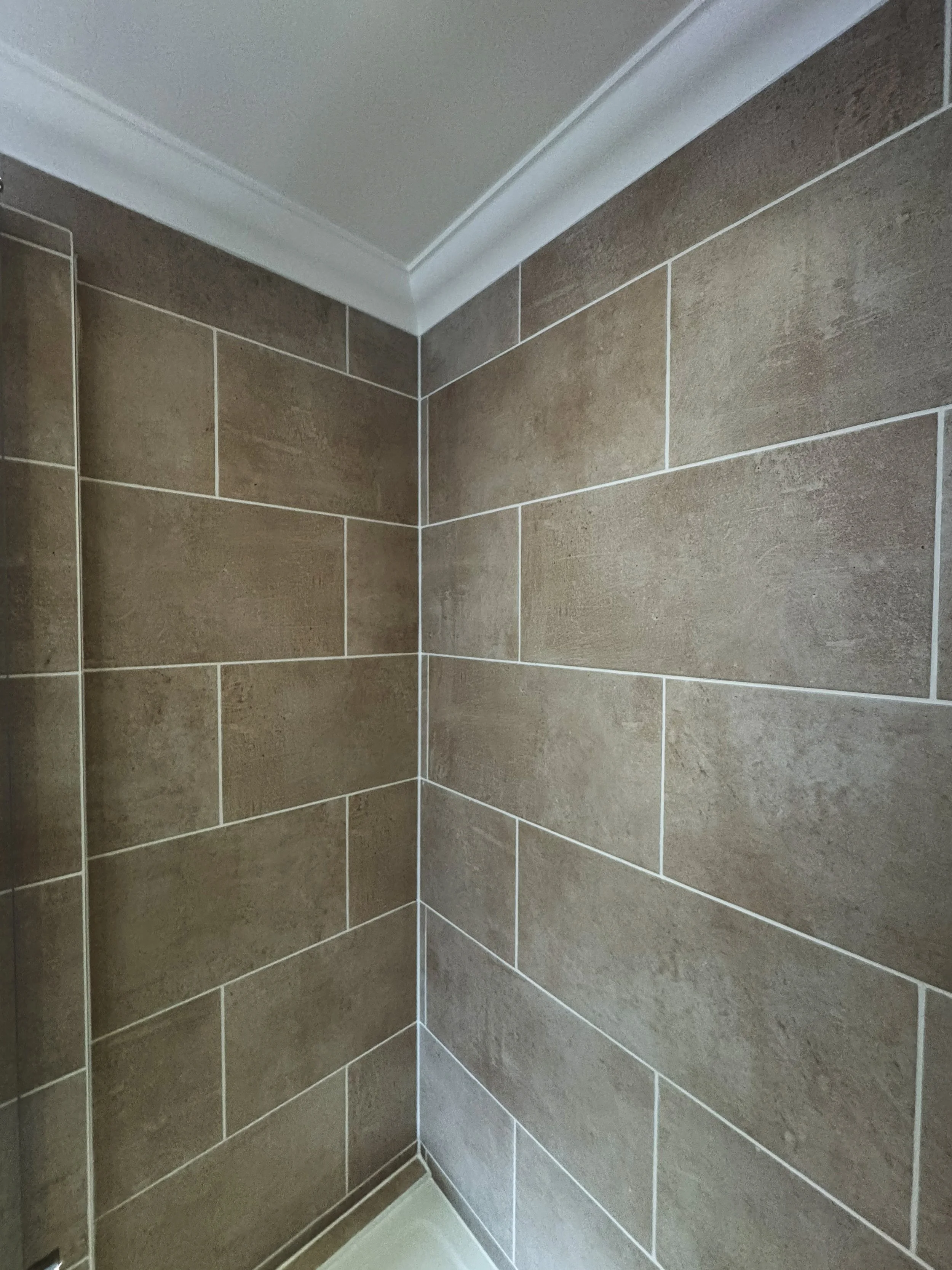 Corner of a shower with brown ceramic tiles and white grout, a white ceiling, and white crown molding.