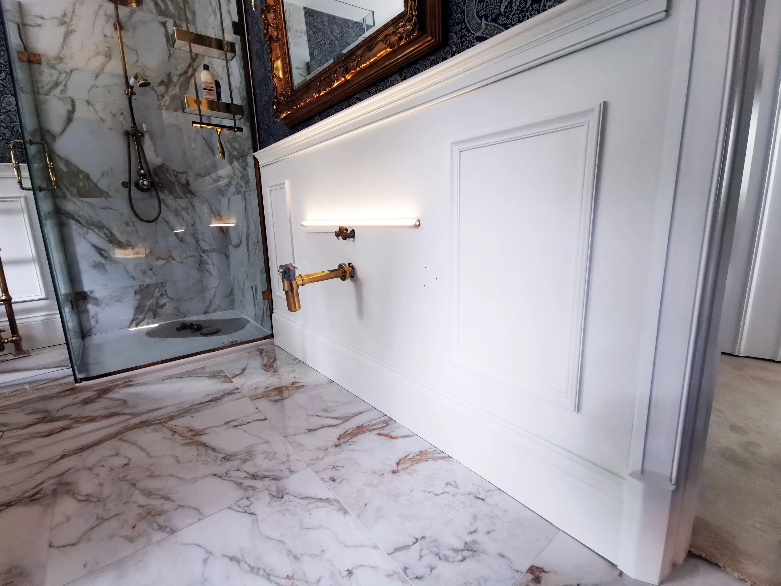 Close-up of a bathroom vanity with a white paneled front, a gold faucet, and a brass-mounted bright strip light above. A framed mirror hangs above the vanity. To the left is a marble walk-in shower with a glass door, marble walls, mounted shower head