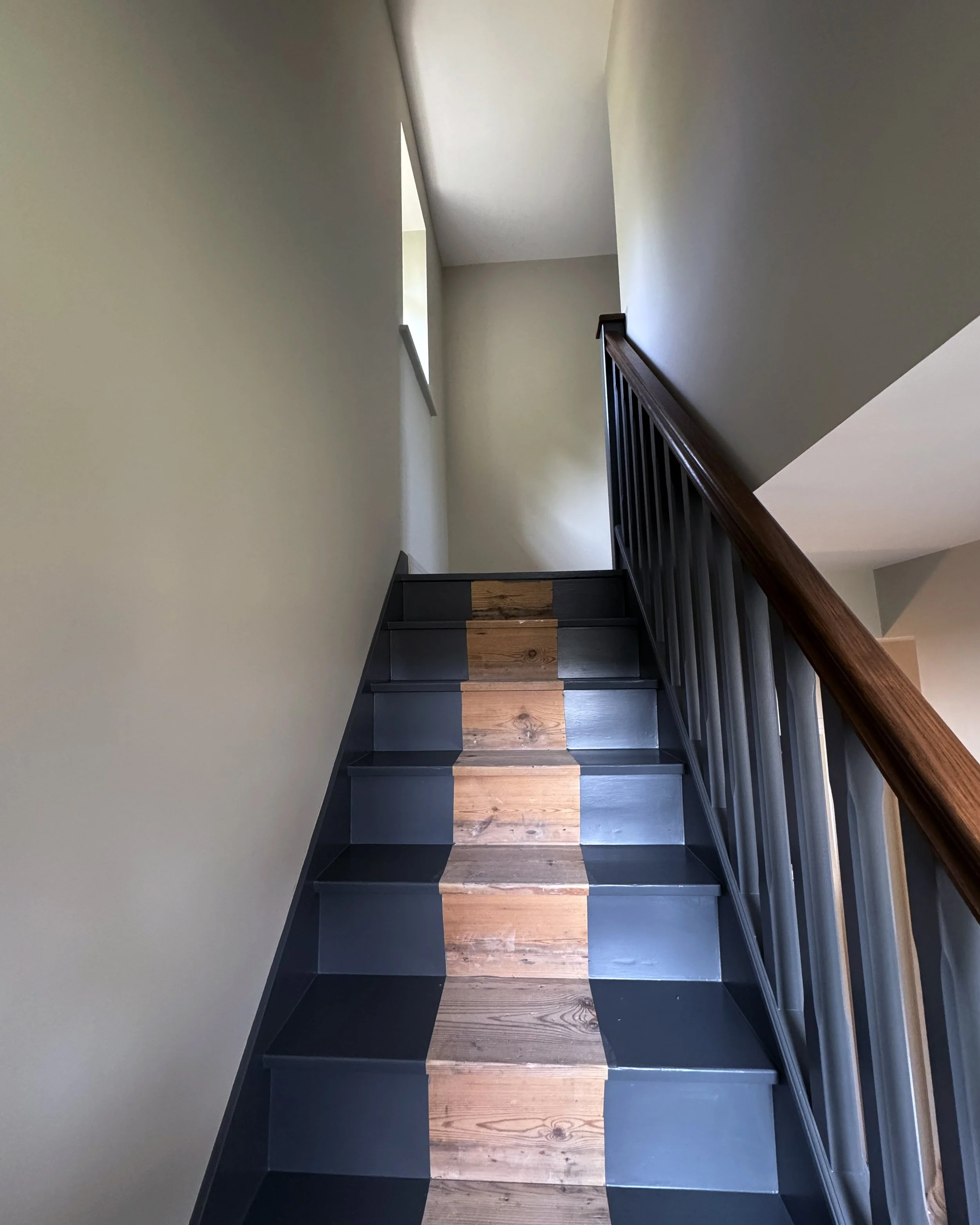 Interior view of a staircase with wooden steps and black risers, a wooden handrail on the right, and a small window at the top letting in natural light.