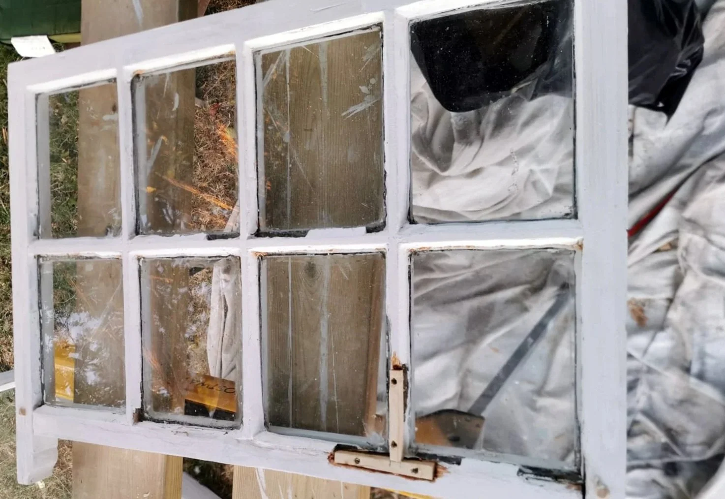 An old wooden and glass window frame with nine panes, some panes are dirty and foggy, painted white, set on a work surface in a workshop or shed, with a person wearing gloves and a apron visible in the background.