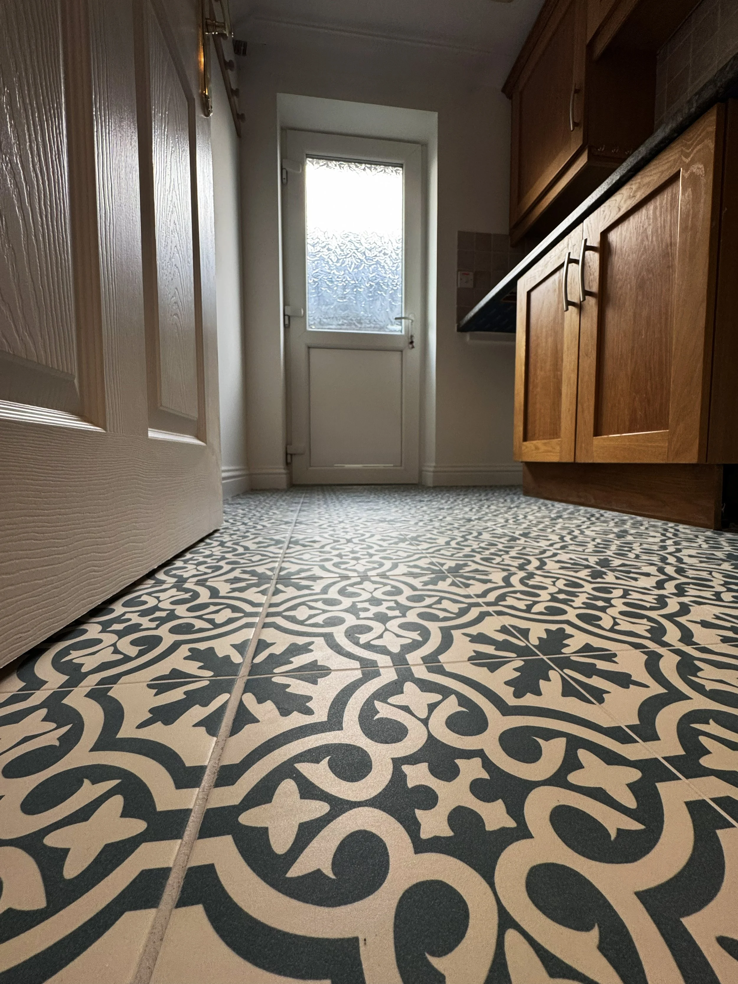 Patterned black and beige tiled floor in a kitchen with wooden cabinets and a door with frosted glass window.
