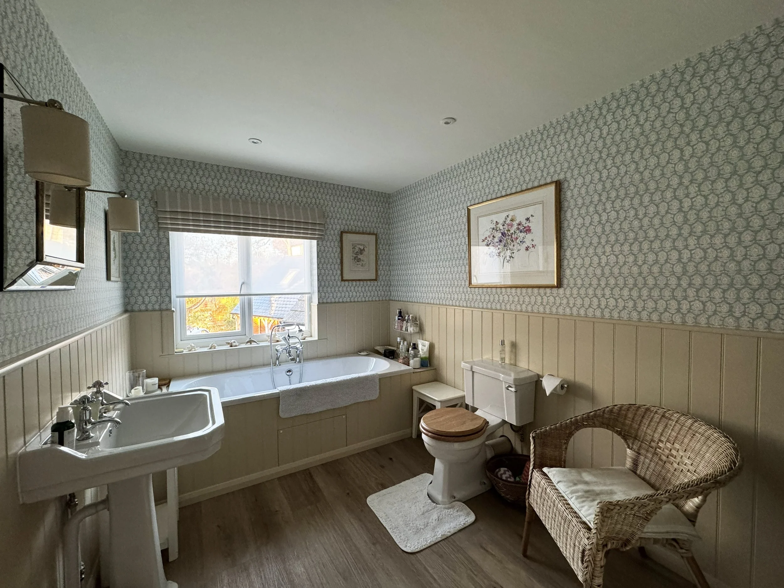 Spacious bathroom with a bathtub, double sink, toilet with wooden seat, wicker chair, framed artwork, and window with patterned shades