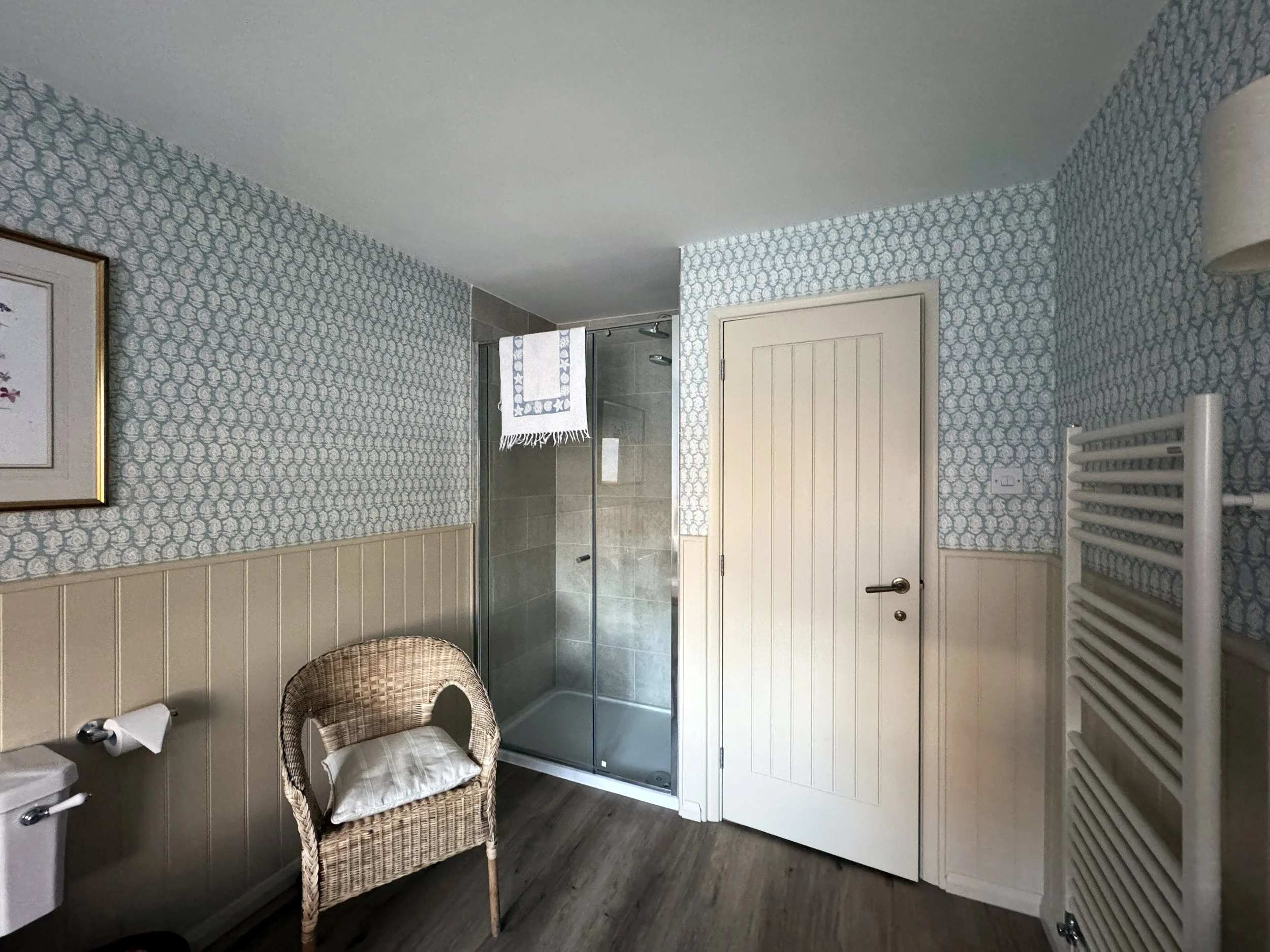 Bathroom with patterned wallpaper, a framed artwork on the wall, a wicker chair with a pillow, a shower stall with glass door, a towel hanging inside, closed white door, a white heated towel rack on the wall, and a wooden floor.
