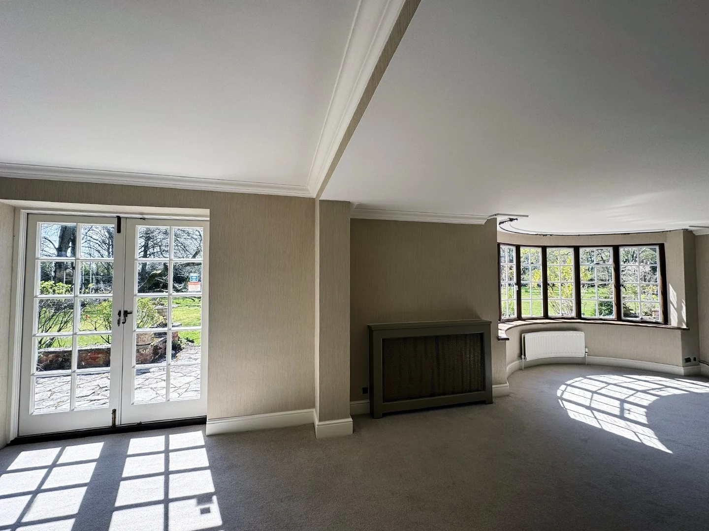 Bright living room with large bay window and French doors leading outside, carpeted floor, and radiator.