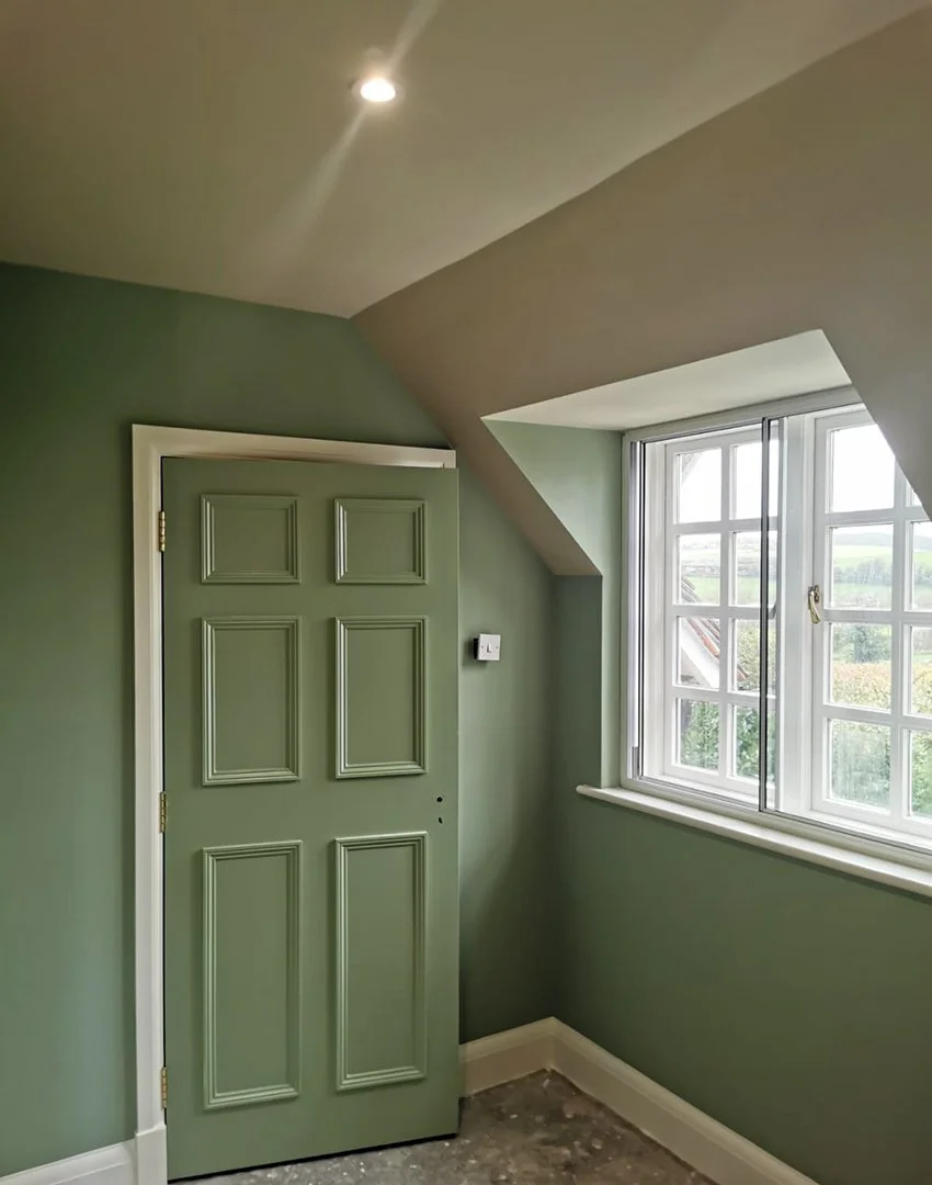 Green interior room with a paneled door, a large window with multiple small panes, and a sloped ceiling.