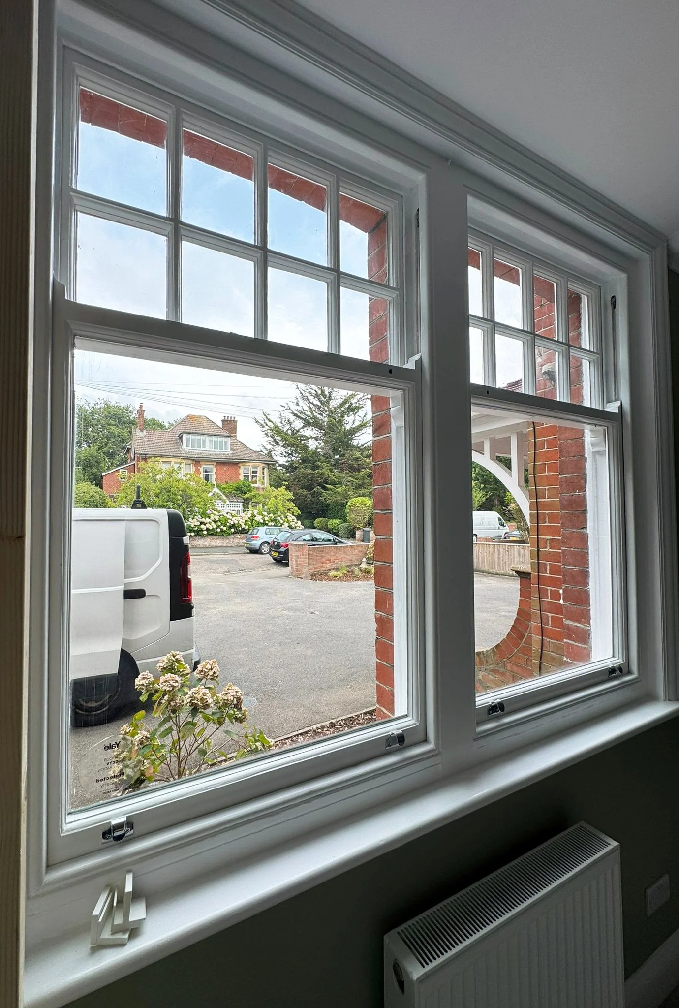 View through a white-framed double-hung window showing a parking lot, a white van, a brick house with trees and bushes, and a clear sky outside.