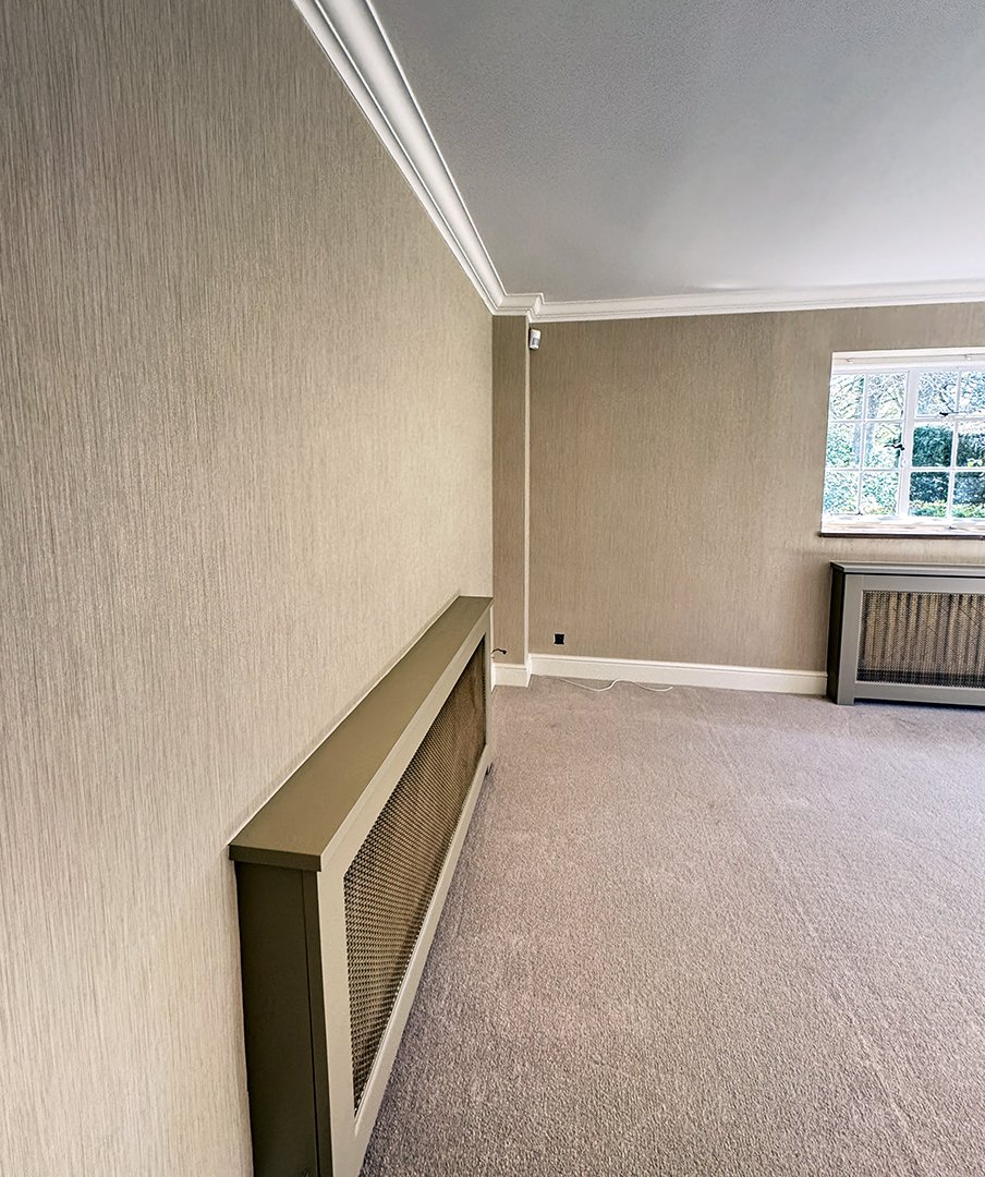Empty room with beige walls, cream carpet, a window showing greenery outside, and a beige radiator cover.