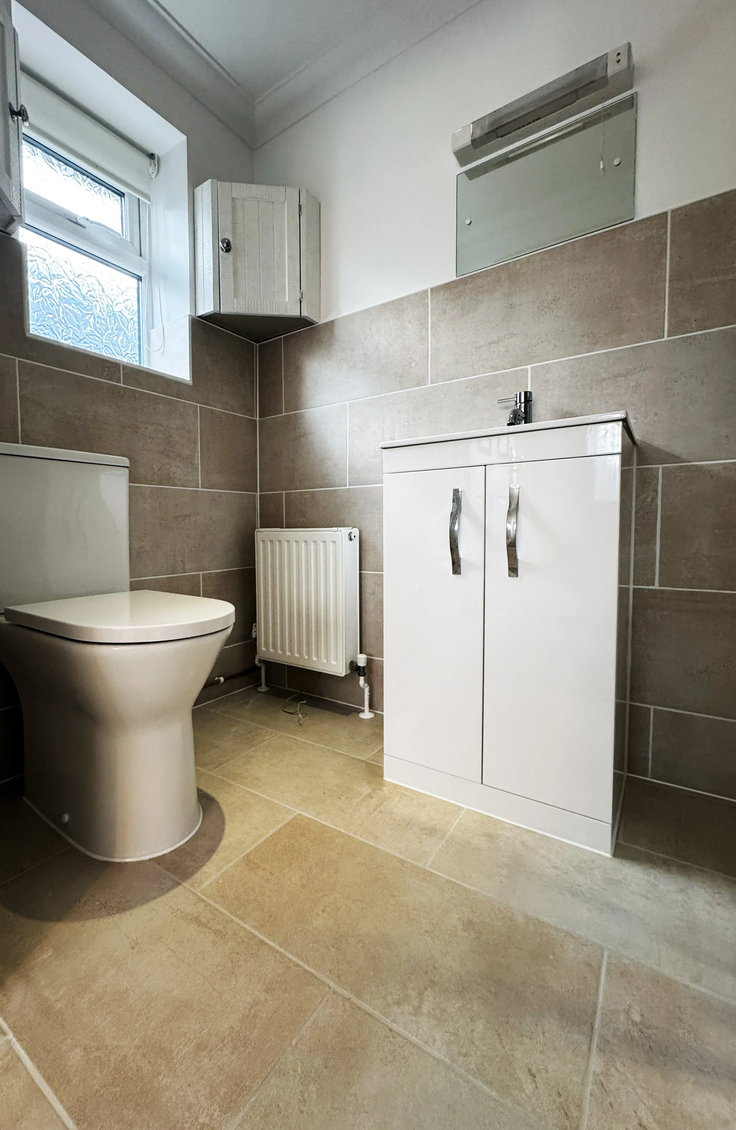 A small bathroom with beige tiled walls and floor, a white toilet, a white bathroom sink cabinet, a small window with frosted glass, a wall-mounted radiator, a small wall cabinet, and a mirror above the sink with a light fixture.