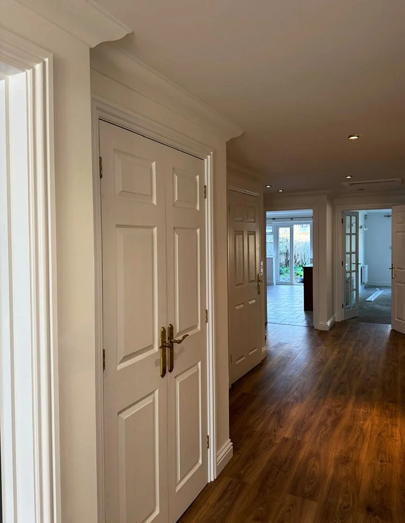 Interior view of a house hallway with multiple closed white doors, hardwood floor, and sliding glass door leading outside.