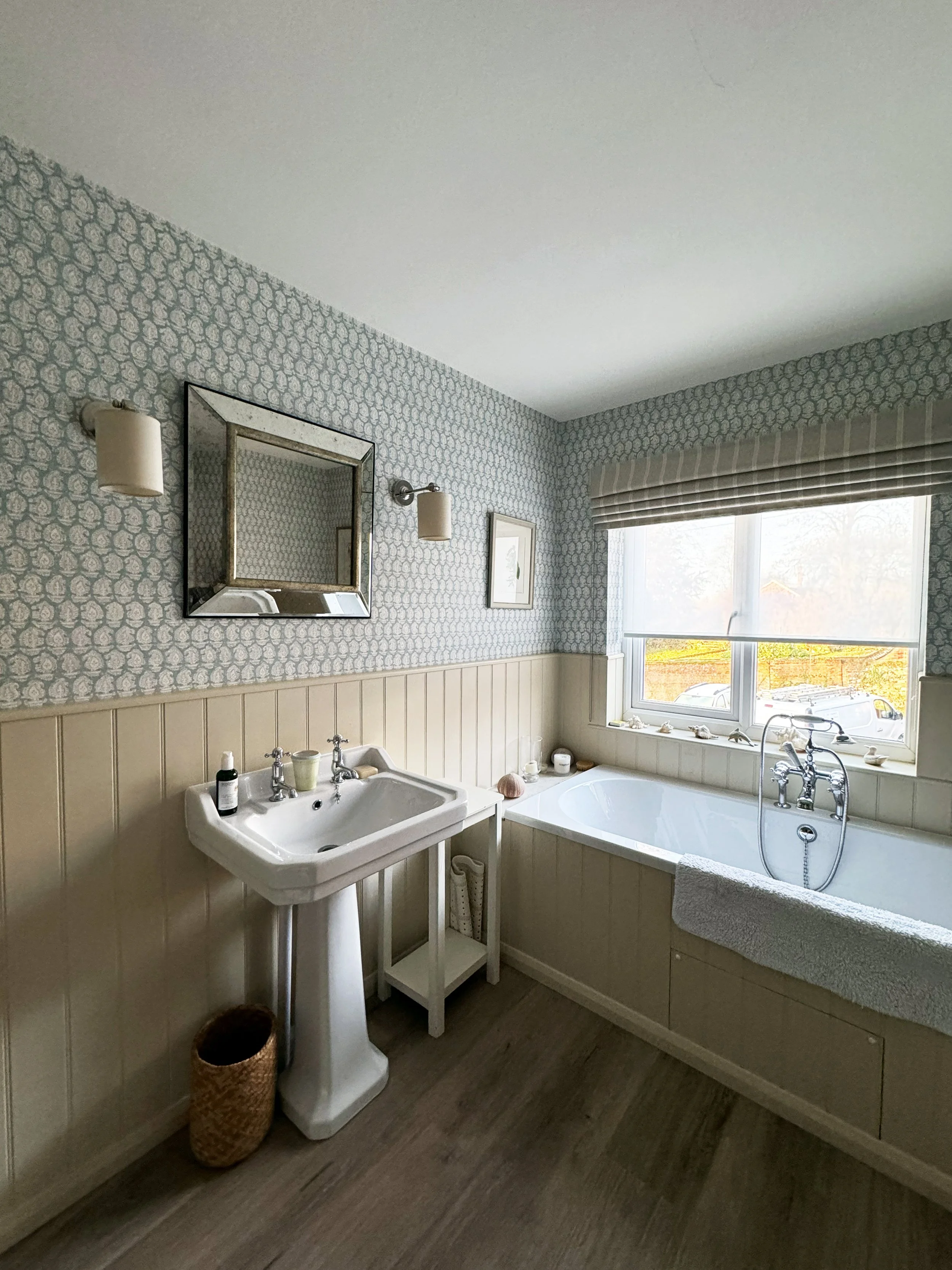 A bathroom with a vintage sink, a bathtub near a window, patterned wallpaper, a mirror, and decorative items on the windowsill.