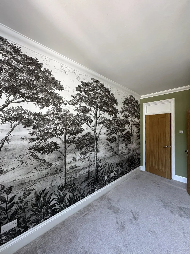 Room with a wallpaper mural depicting a black and white landscape scene with trees, mountains, and plants. The room has a beige carpet, a wooden door, and a small portion of a green painted wall.