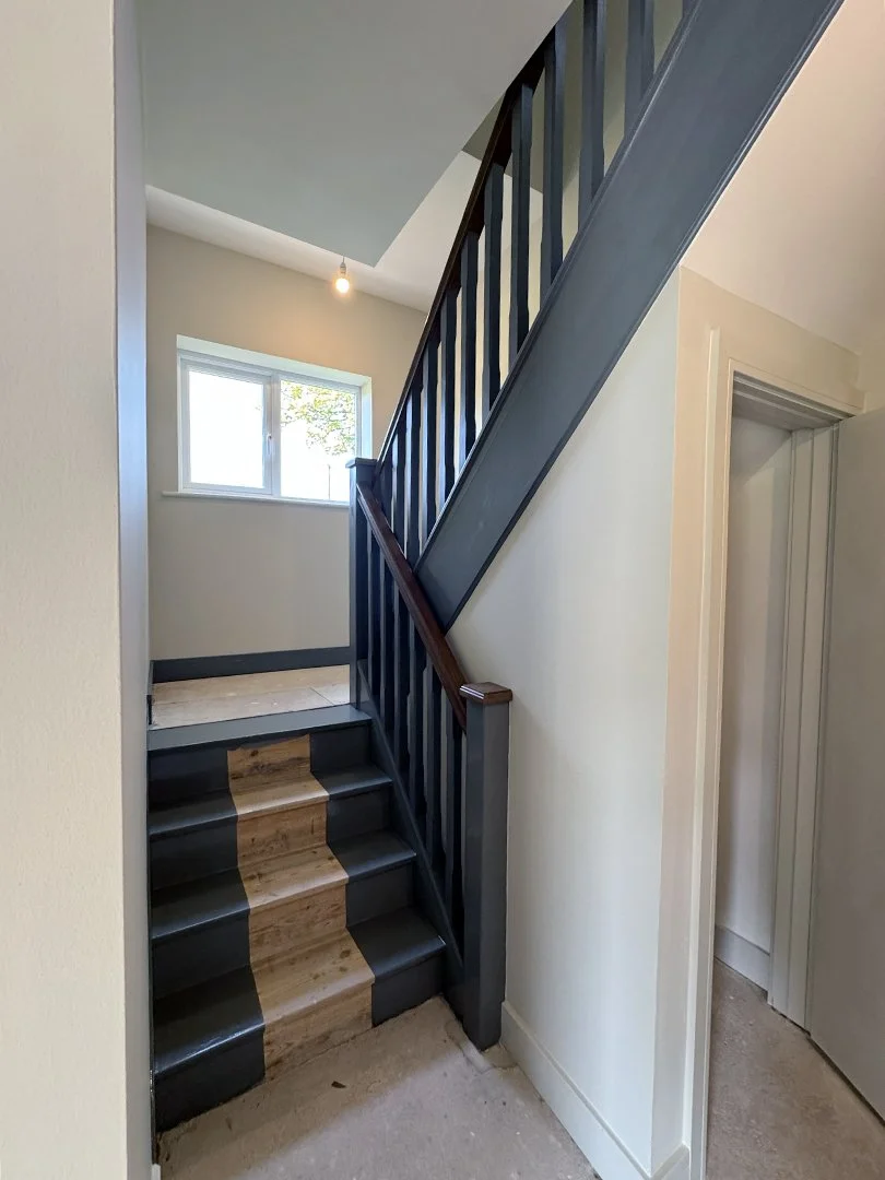 Interior view of a staircase with black-painted steps and a wood-colored strip running down the center. The staircase has a black wooden handrail and balusters, leading to a room with a window letting in natural light. The walls are light-colored, an