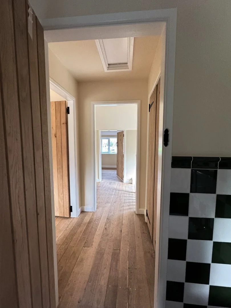 View of a hallway in a house with wooden floors, beige walls, and multiple rooms with wooden doors. There is a skylight in the ceiling and a window in the room at the end of the hallway.