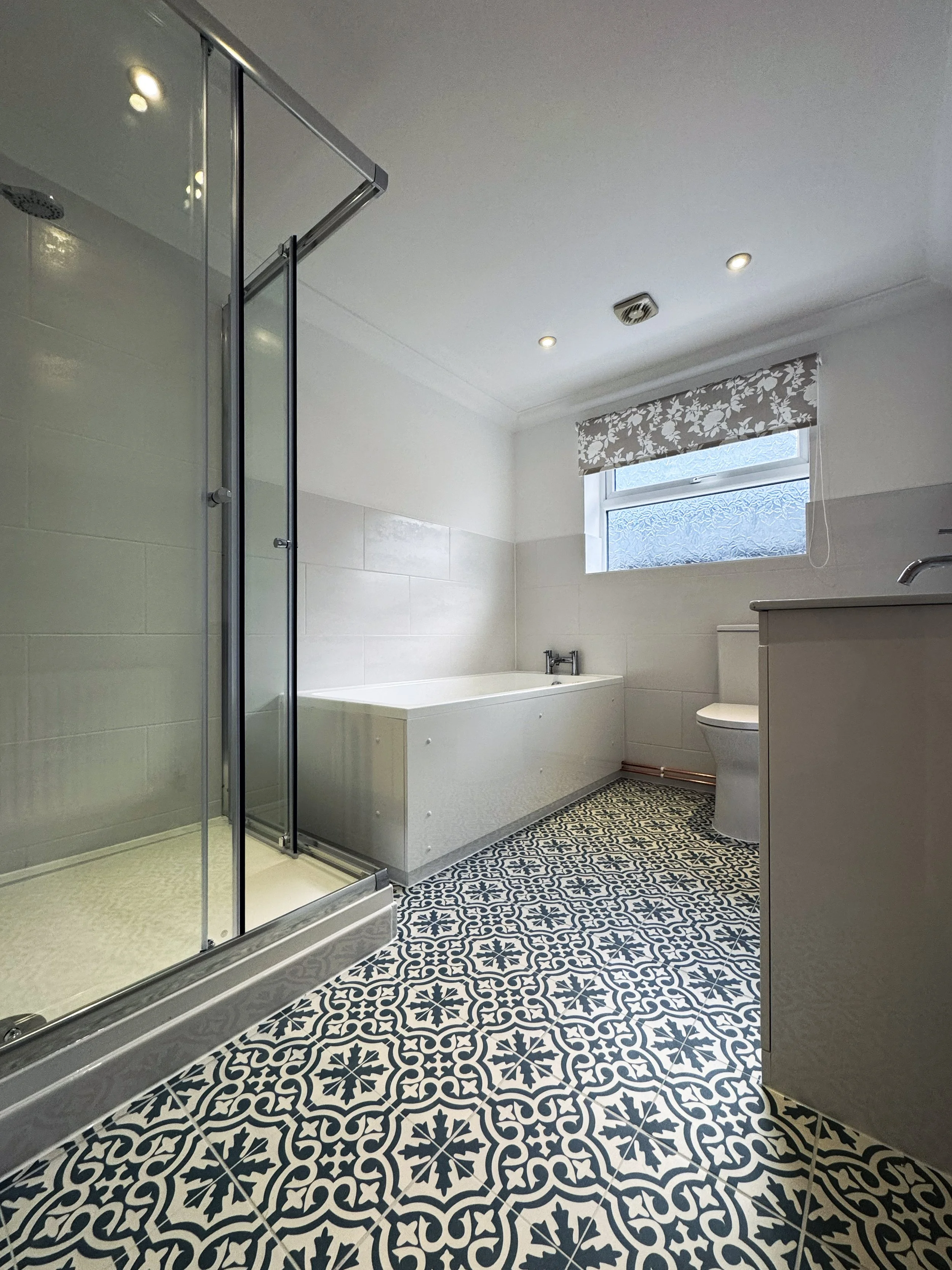 Bathroom with patterned black and white tiled floor, shower stall, bathtub, toilet, and vanity, with a small window and floral curtain.