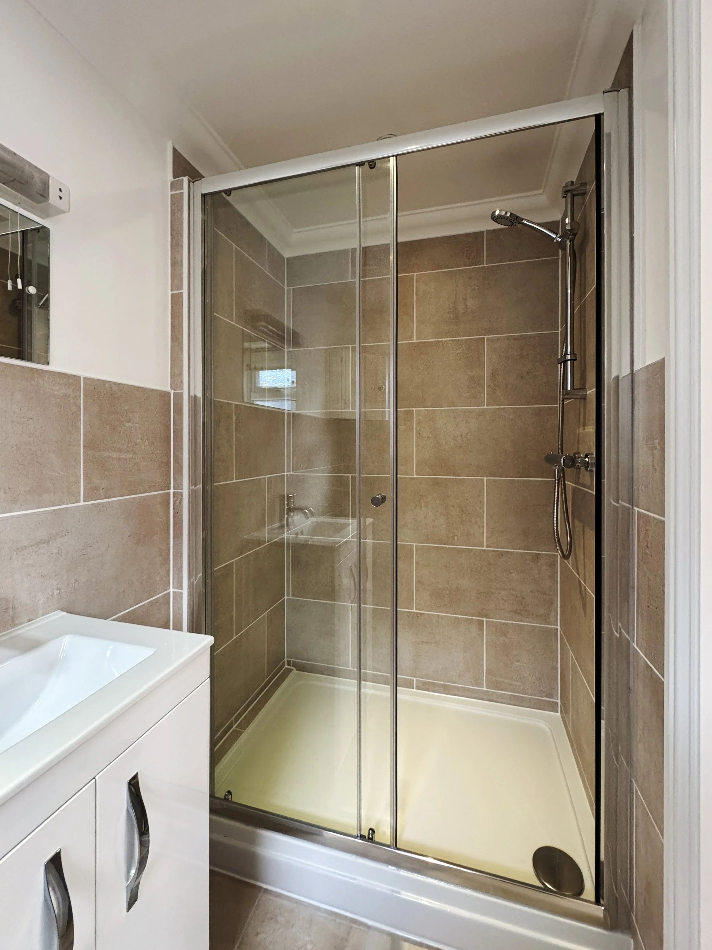 Bathroom shower with glass doors, brown tiled walls, and a handheld showerhead, adjacent to a white vanity with a sink.