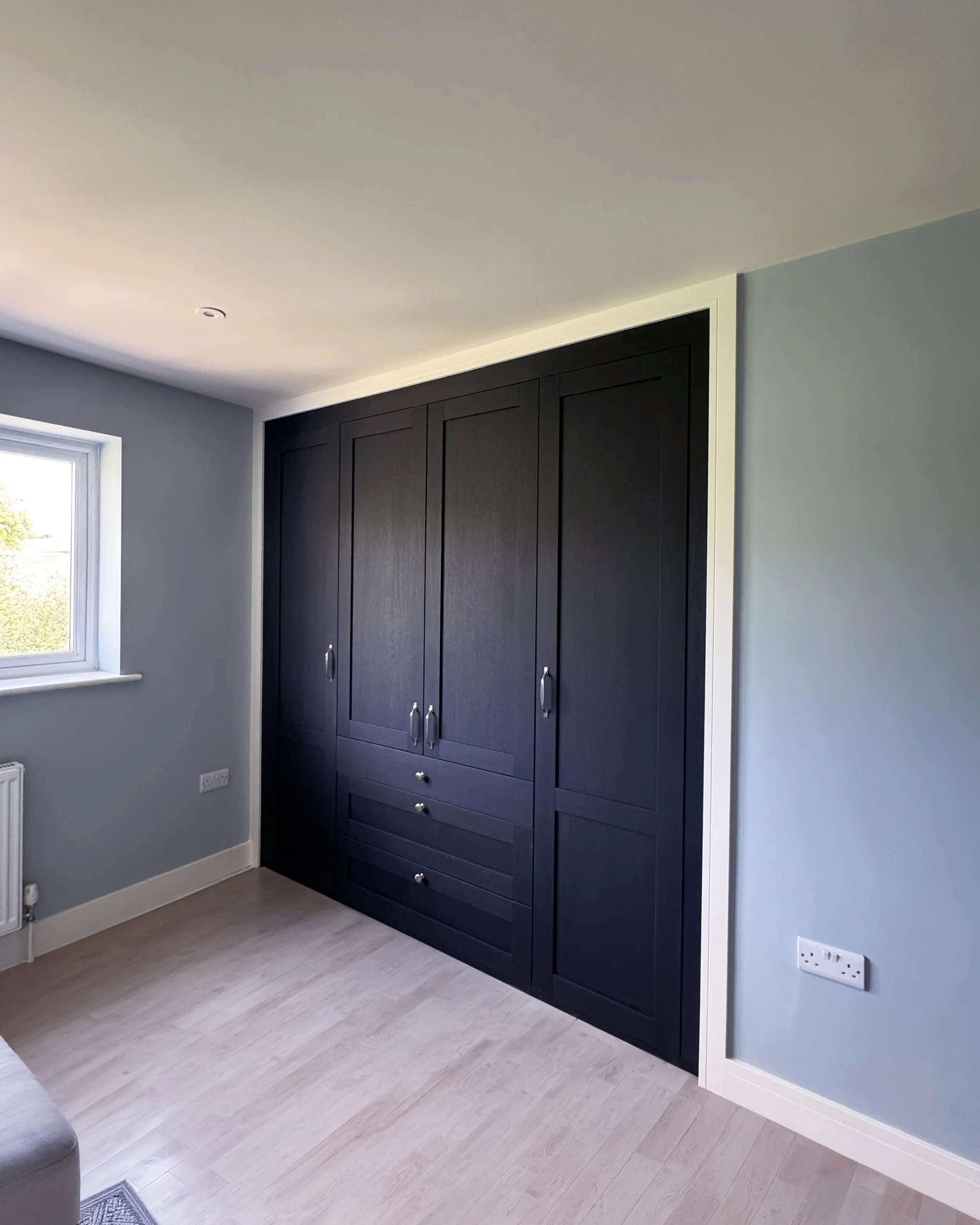Modern bedroom with a black wardrobe, light blue walls, a small window, and a light-colored wooden floor.