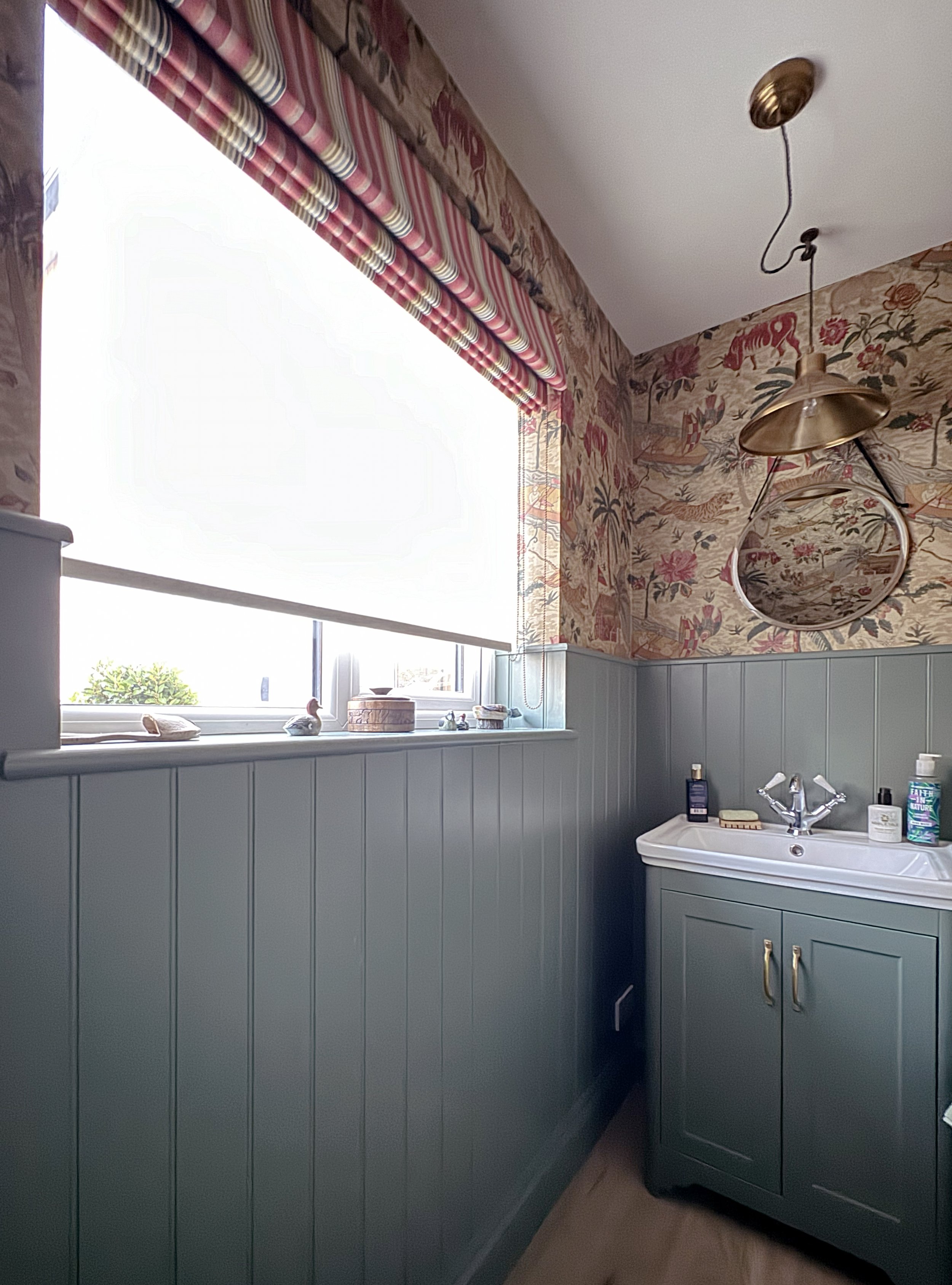 A vintage-style bathroom with a window covered by a white blind, pastel green wainscoting, and floral wallpaper. There is a small sink with a mirror above, and decorative items and toiletries on the window ledge.