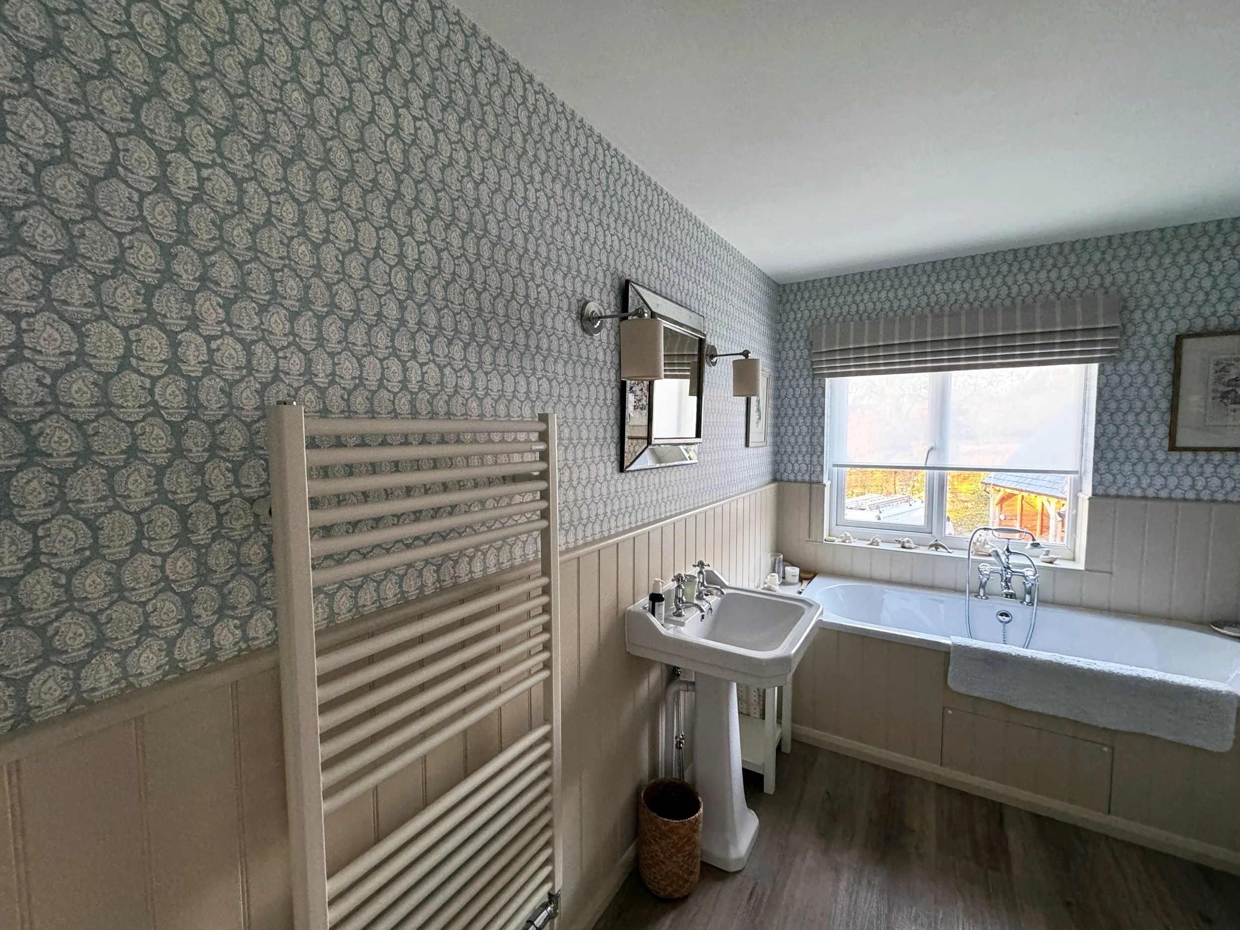 A bathroom with a patterned wallpaper, a window with blinds, a bathtub, a sink, a mirror, a wall lamp, and a radiator.