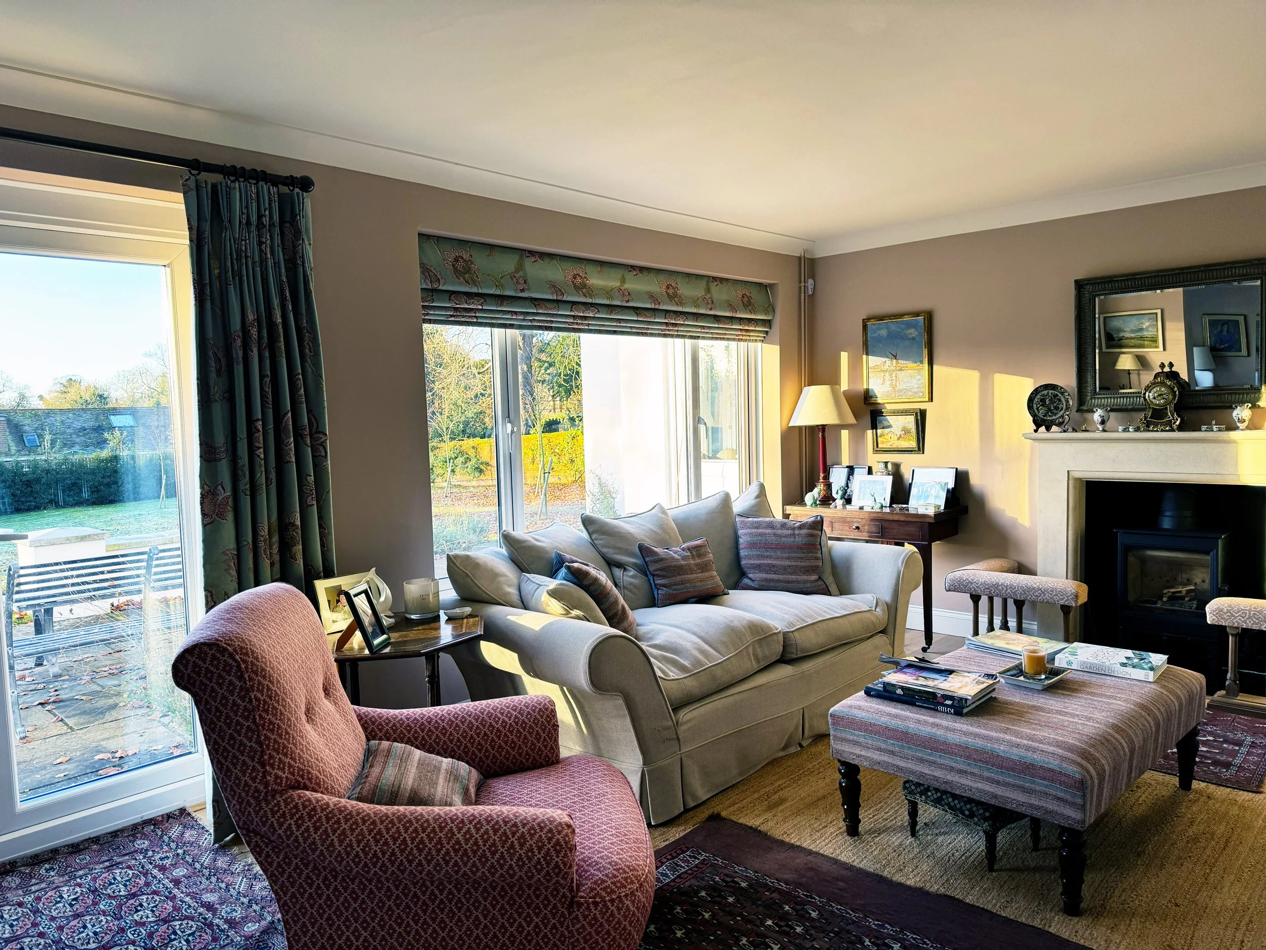 Living room with cream sofa, purple armchair, striped ottoman, fireplace, wall mirror, colorful cushions, side table, window with floral blinds, and garden view outside.