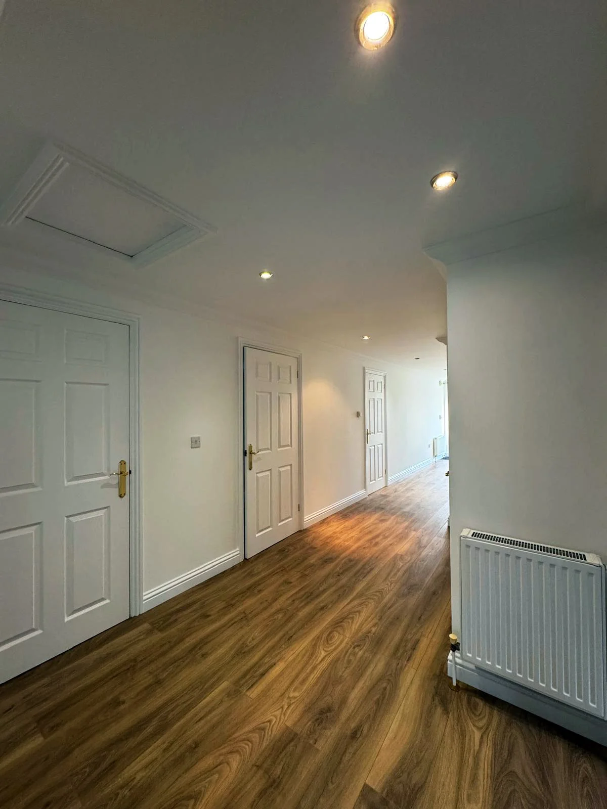 Empty hallway with white walls, wooden flooring, and closed white doors, illuminated by recessed ceiling lights, and a white radiator on the right side.