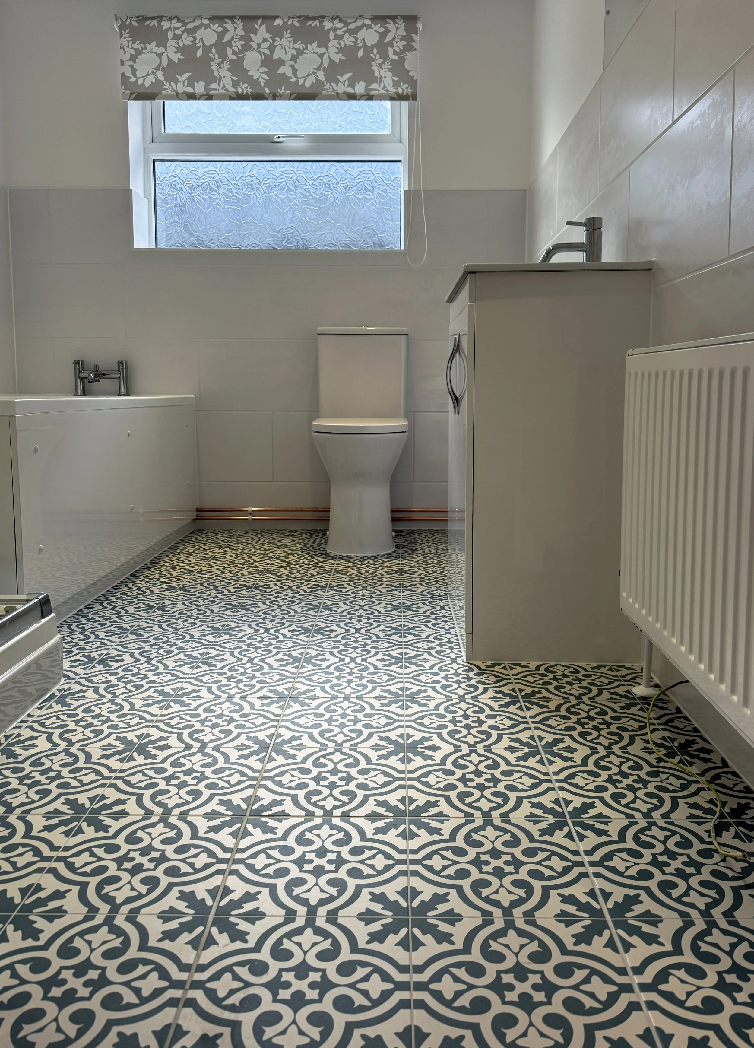 A clean bathroom with patterned black and white floor tiles, a white toilet, a window with frosted glass and floral curtains, a white radiator, and a white vanity with a sink.