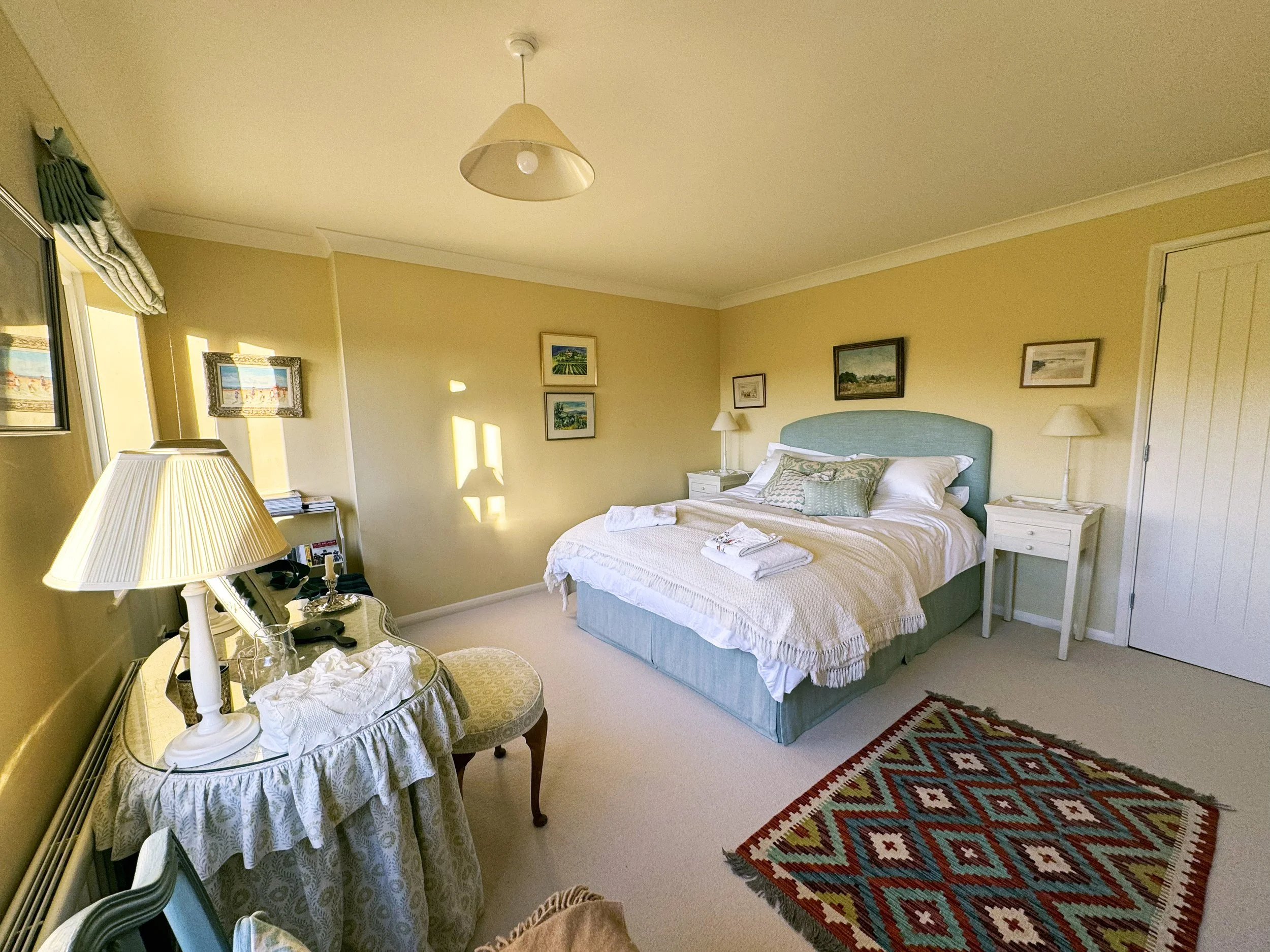 A cozy bedroom with a large bed featuring a light blue headboard, white bedding, and decorative pillows. Beside the bed are matching white nightstands with lamps. The room has yellow walls adorned with framed artwork, a colorful geometric rug on the 