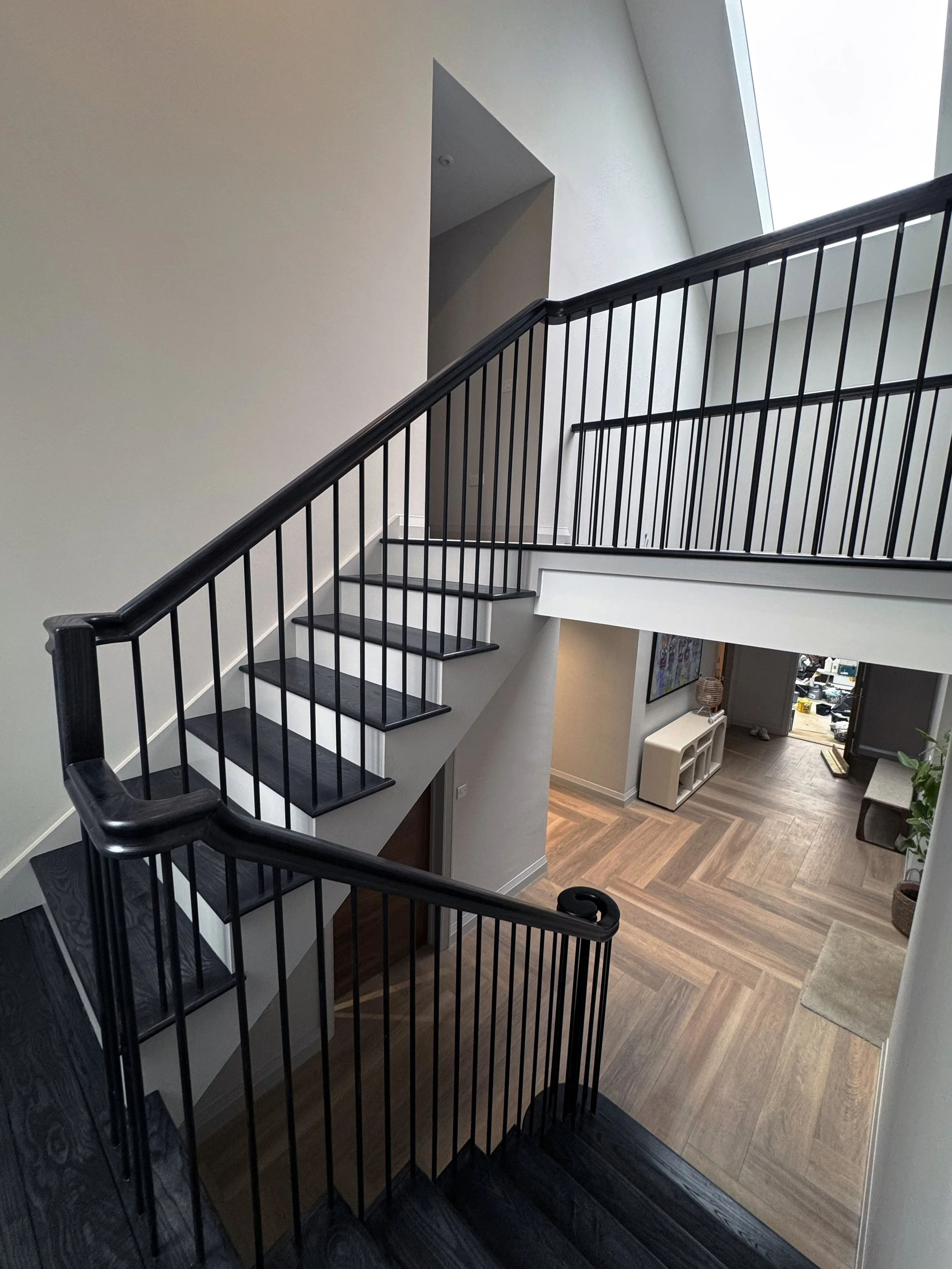 Interior view of a modern home with a black-walled staircase leading down to a living space with wooden flooring, a white cabinet, and a doorway, illuminated by natural light from a skylight.