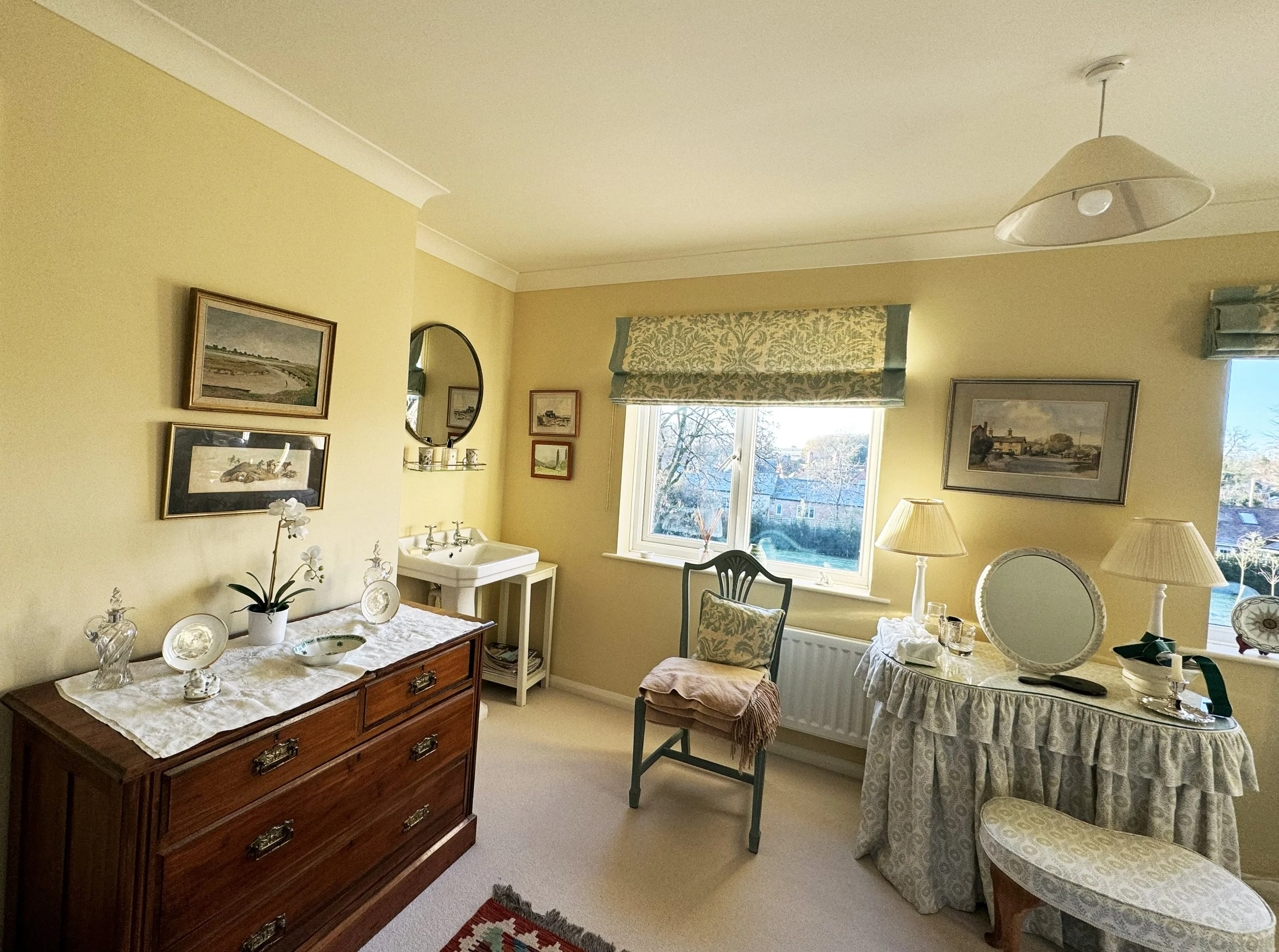 A cozy, well-decorated living room with yellow walls, featuring a wooden dresser with decorative items, a small sink, framed pictures on the wall, a window with a patterned curtain, a green chair with a cushion and a pink throw, a oval mirror, two ta