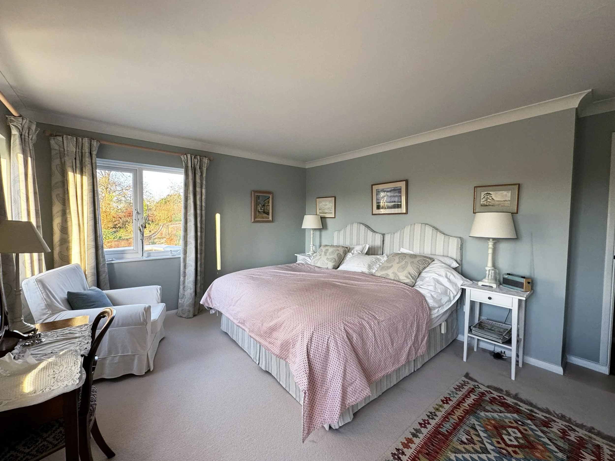 A bedroom with a large bed covered in pink bedding, two bedside lamps, a white armchair with a pillow, and a window with curtains showing a view of trees outside.