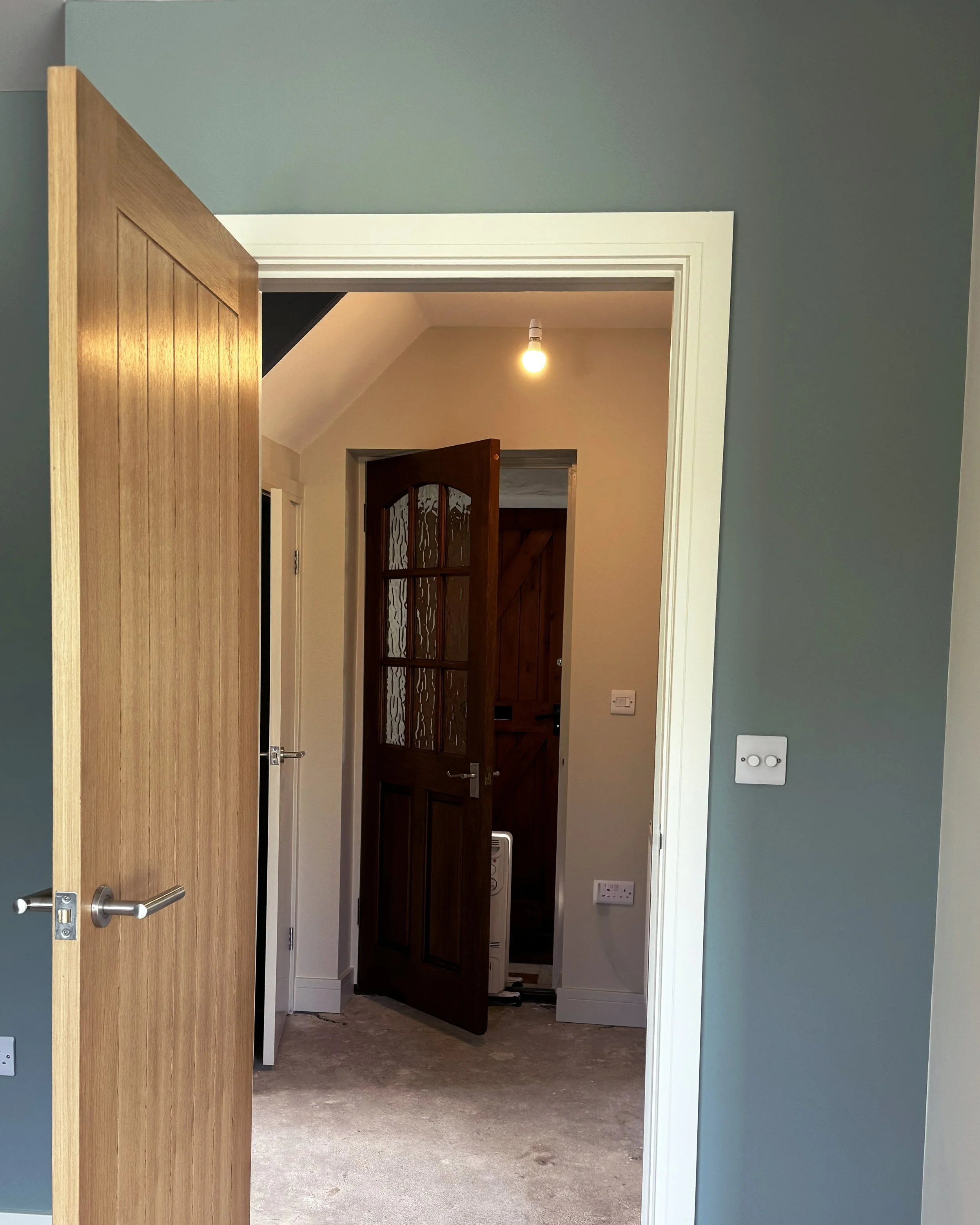View through an open door into a small room with a second partially open door, with a hanging lightbulb and beige walls.