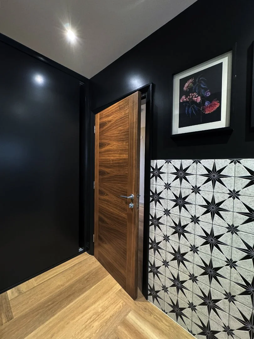 Interior corner with a wooden door, black walls, patterned ceramic tiles on the wall, and framed artwork with a floral design.