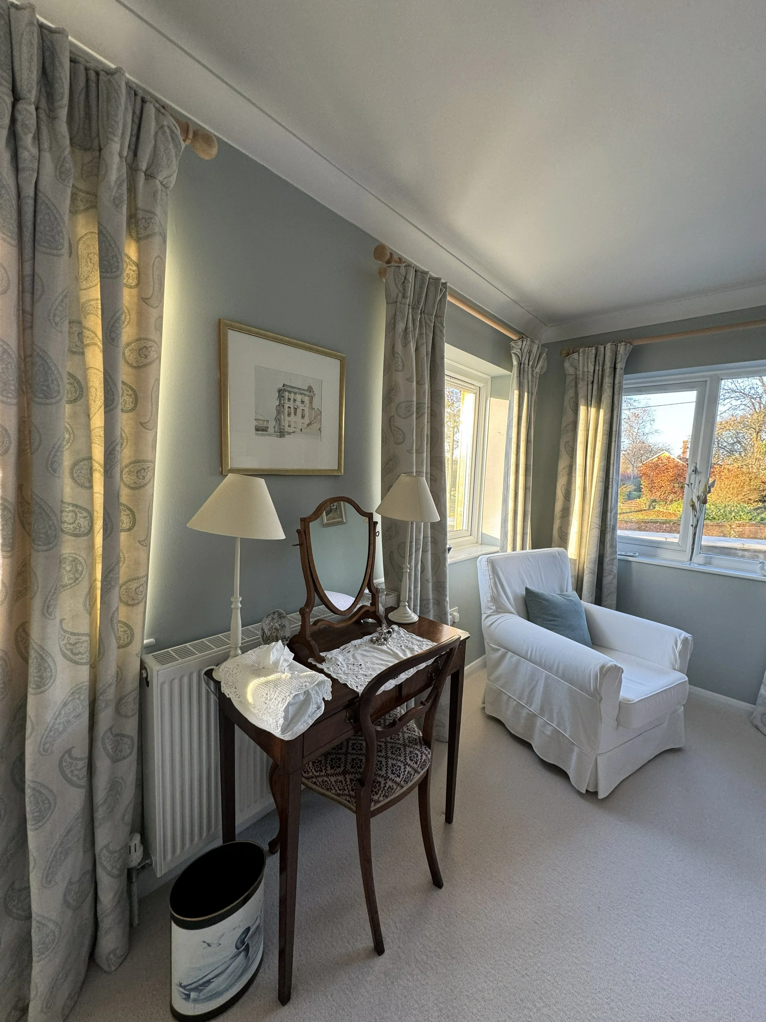 A corner of a cozy living room with a white armchair, a wooden vanity table with a mirror, lamps, and a framed artwork on a light-colored wall. There are large windows with patterned curtains letting in natural light.