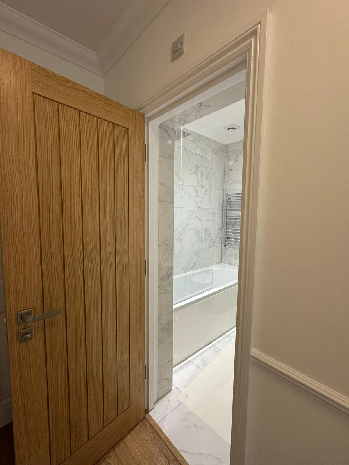 View into a bathroom through an open wooden door, showing marble tiling on the walls and a bathtub.