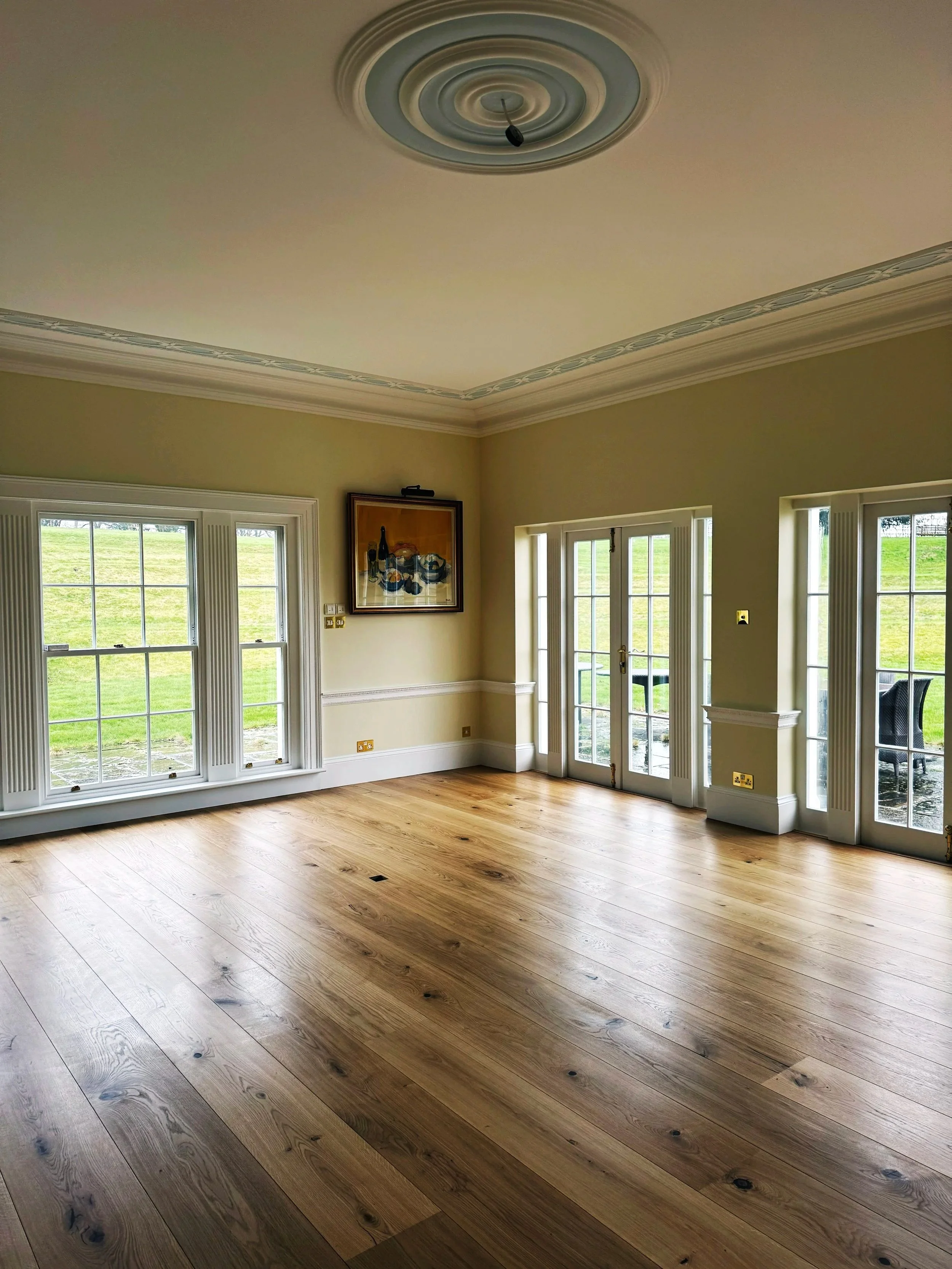 Empty living room with large windows and glass doors, hardwood floors, beige walls, and decorative crown molding.