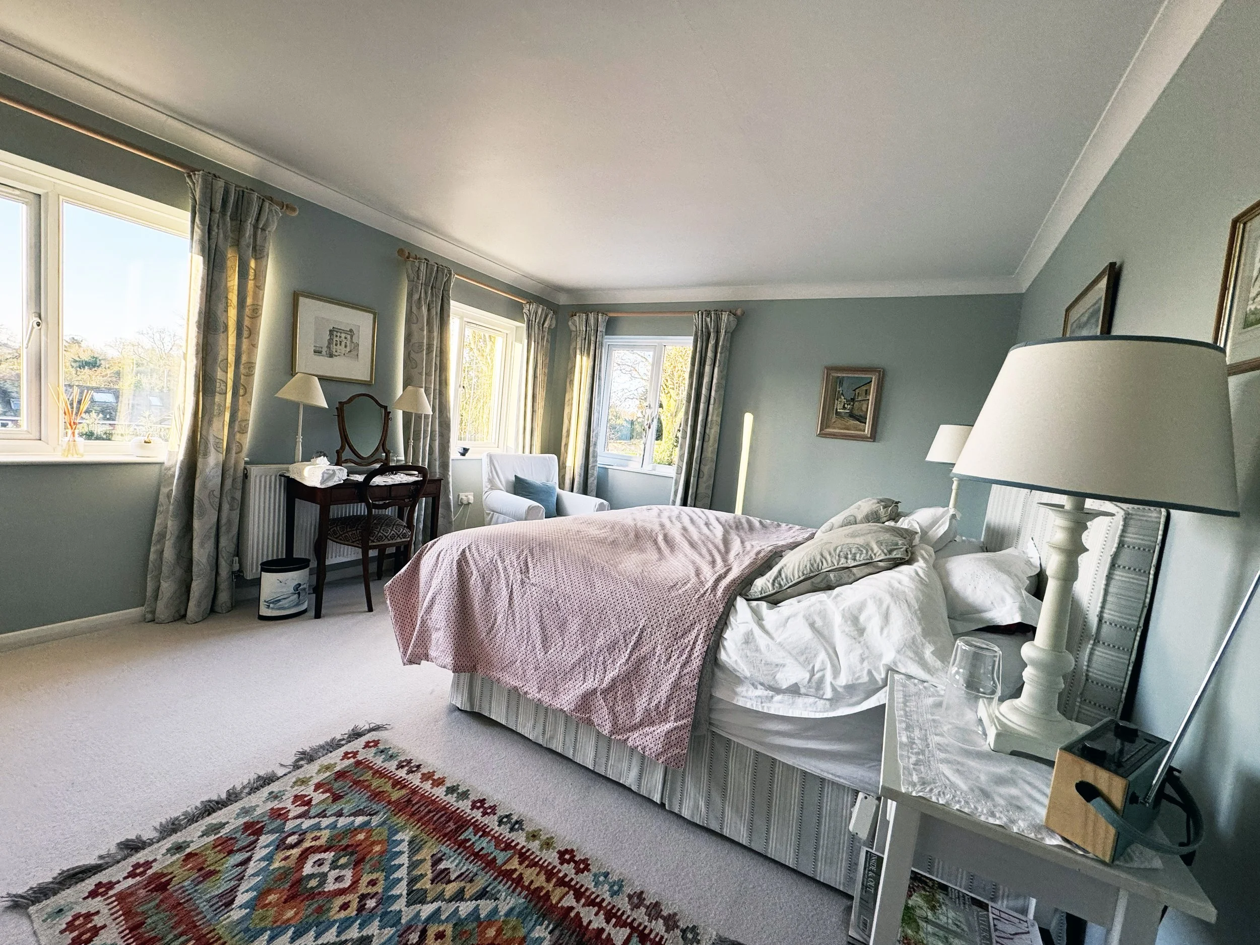 Bright bedroom with a large bed, white bedding, pink blanket, and multiple pillows. Four windows with curtains let in natural light. Traditional furniture includes a side table with a lamp, a dresser with a mirror, and a desk with lamps. Decorated wi