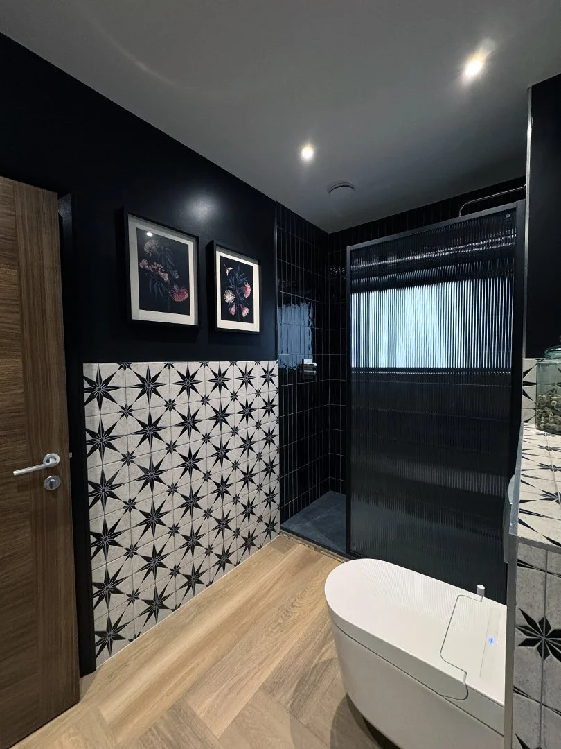 Modern bathroom with black and white geometric tile on one wall, dark tiled shower area, wooden floor, and a wall with framed floral art.