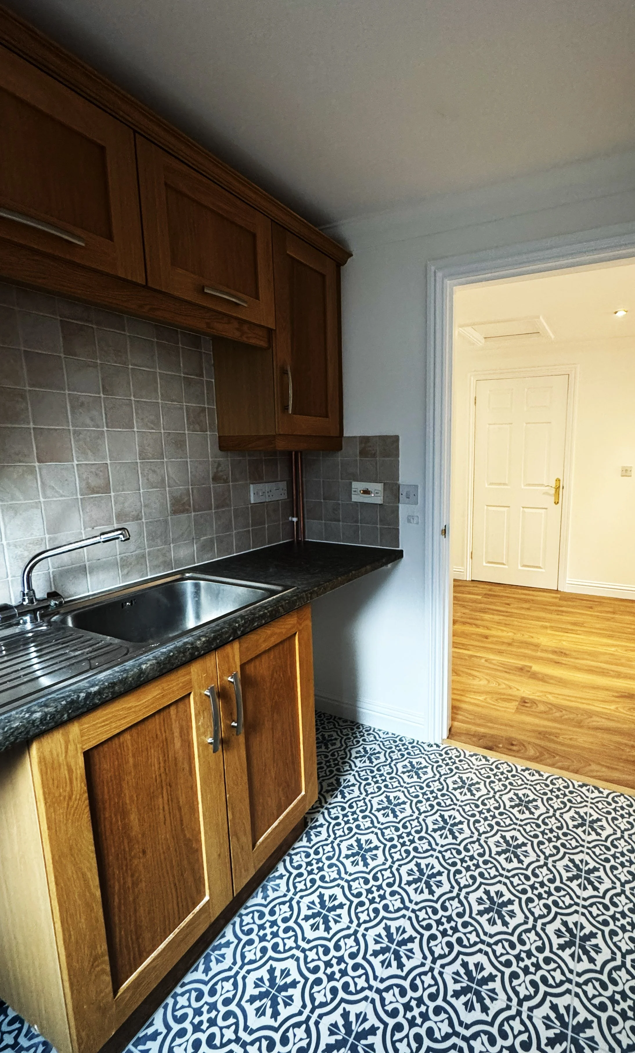 Kitchen with wooden cabinets, black countertop, stainless steel sink, patterned tile floor, and beige tiled backsplash.