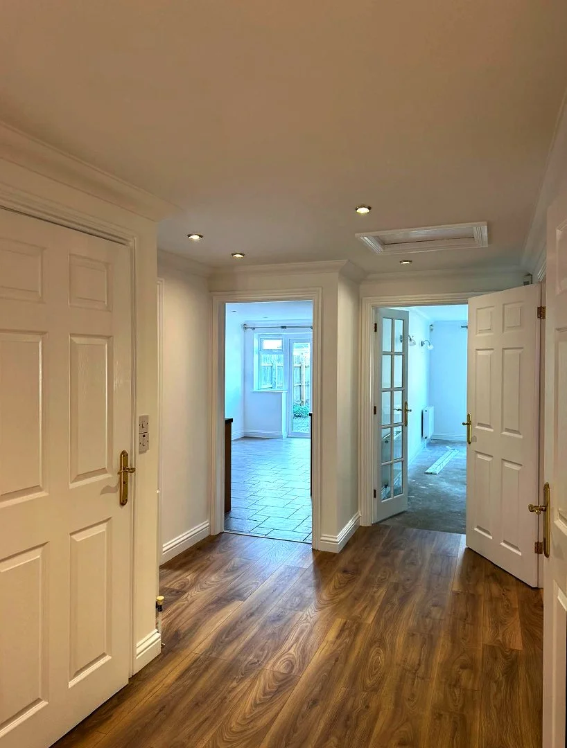 Interior view of a hallway with laminate wood flooring, white walls, and multiple open doors leading to rooms with tiled and carpeted floors. Recessed ceiling lights illuminate the space.