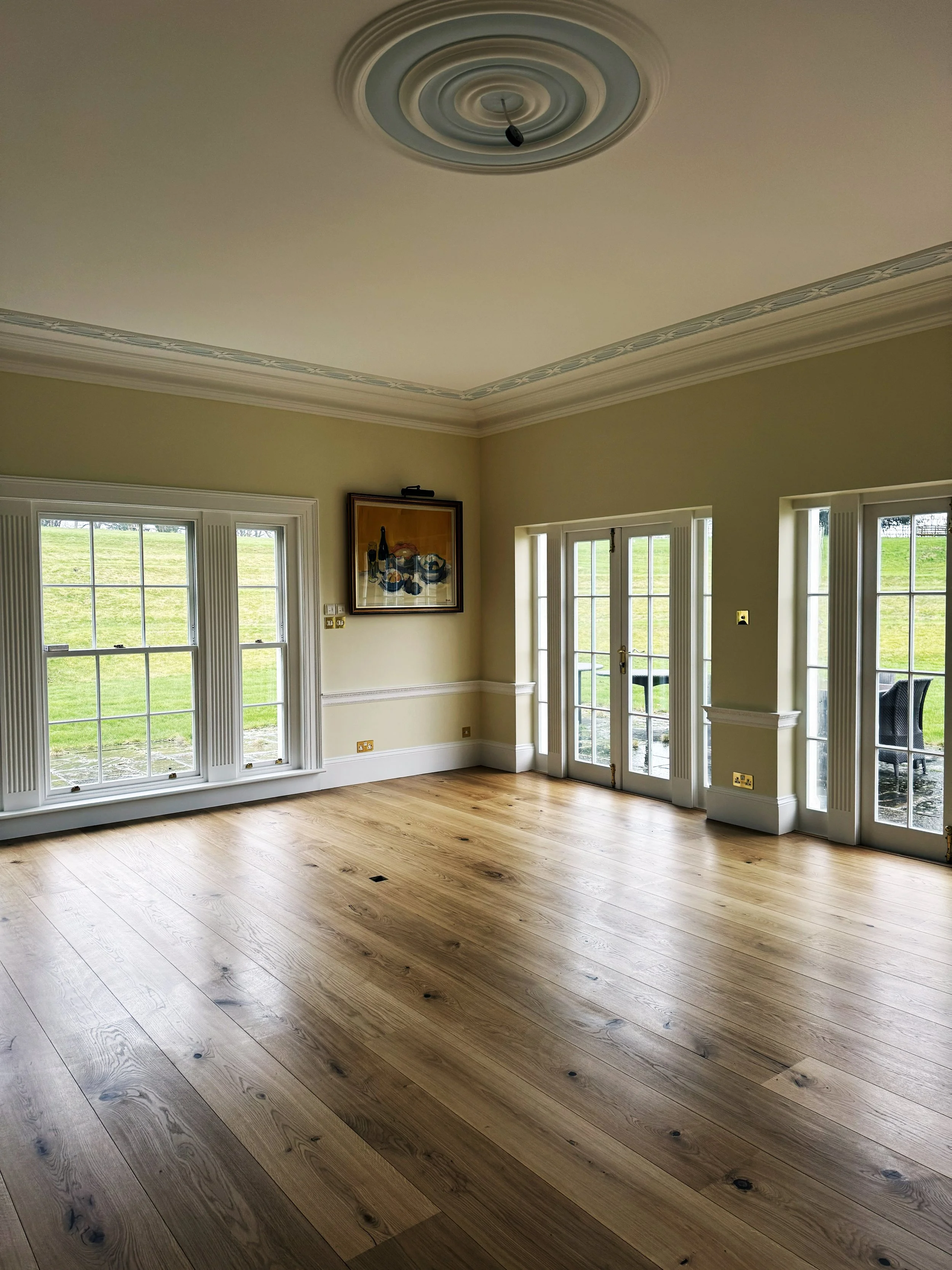 Empty living room with large windows and a glass door leading outside, hardwood flooring, beige walls, and a decorative ceiling with a light fixture. There is a framed painting on the wall.