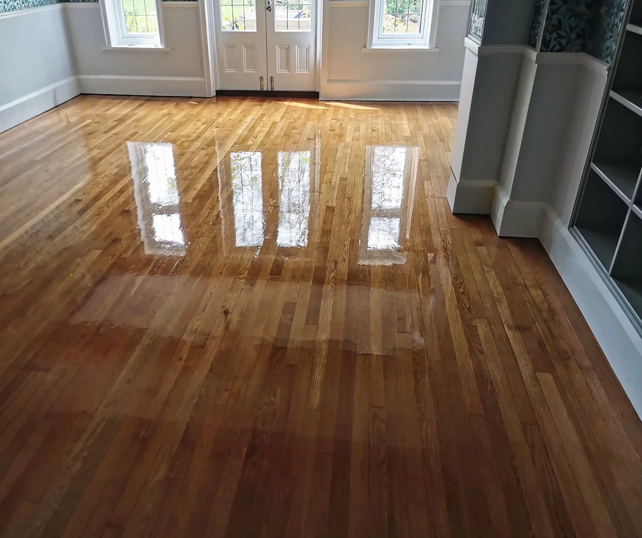 A room with freshly polished hardwood floors reflecting light from two windows and a glass door, with white walls and a built-in shelf on the right.