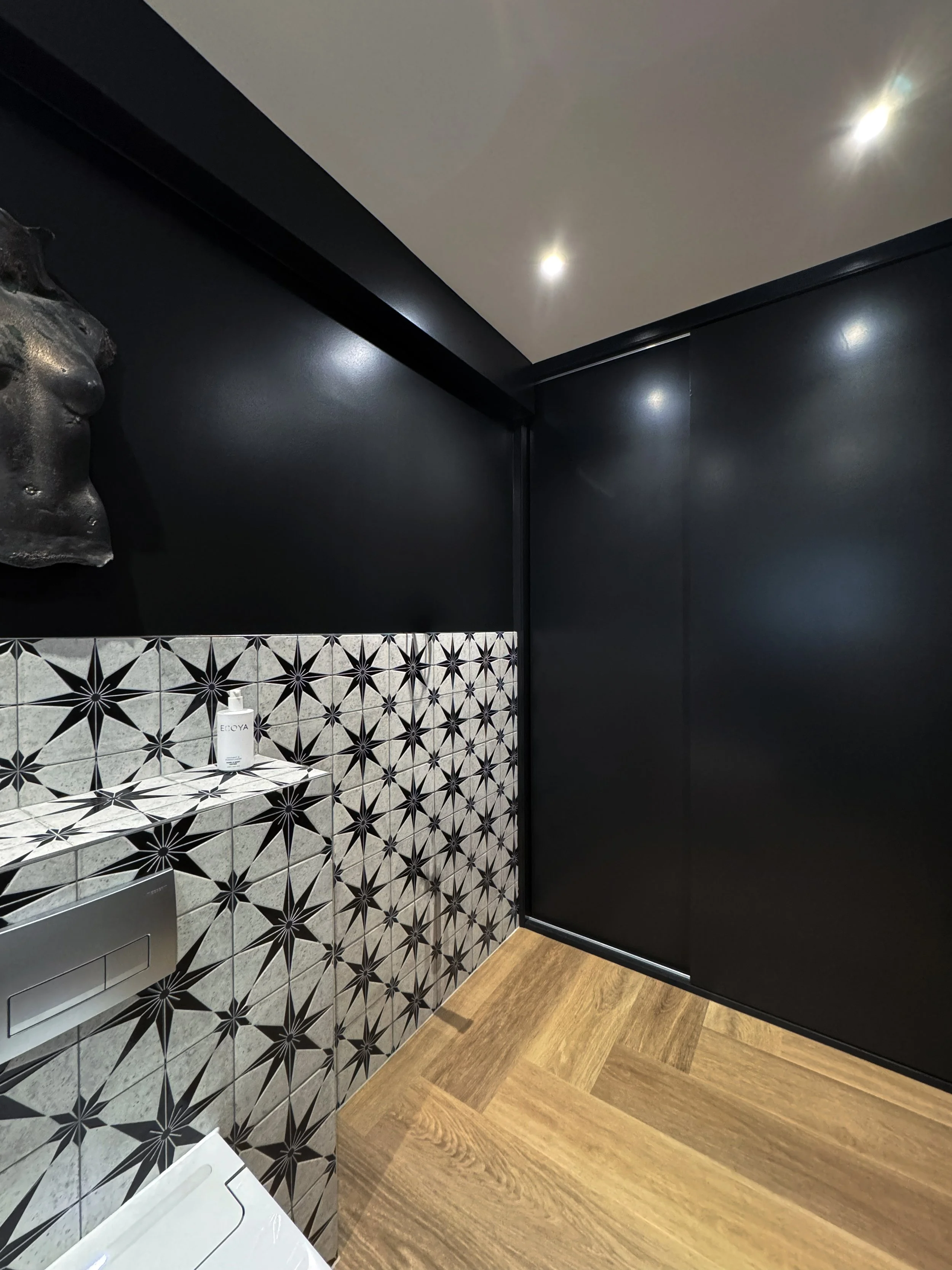 Interior corner of a modern room with black walls, patterned tile backsplash with black star designs, a small shelf with a bottle of hand sanitizer, wood floor, and a white object on the floor.