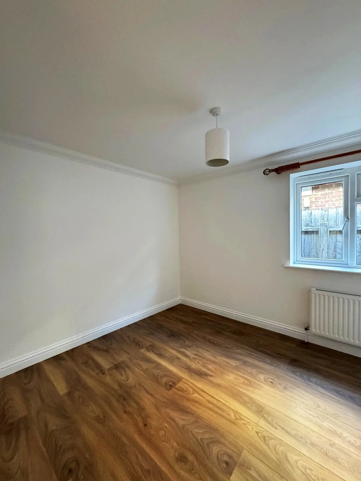 Empty room with white walls, a window with a wooden curtain rod, a radiator below the window, and wood laminate flooring.