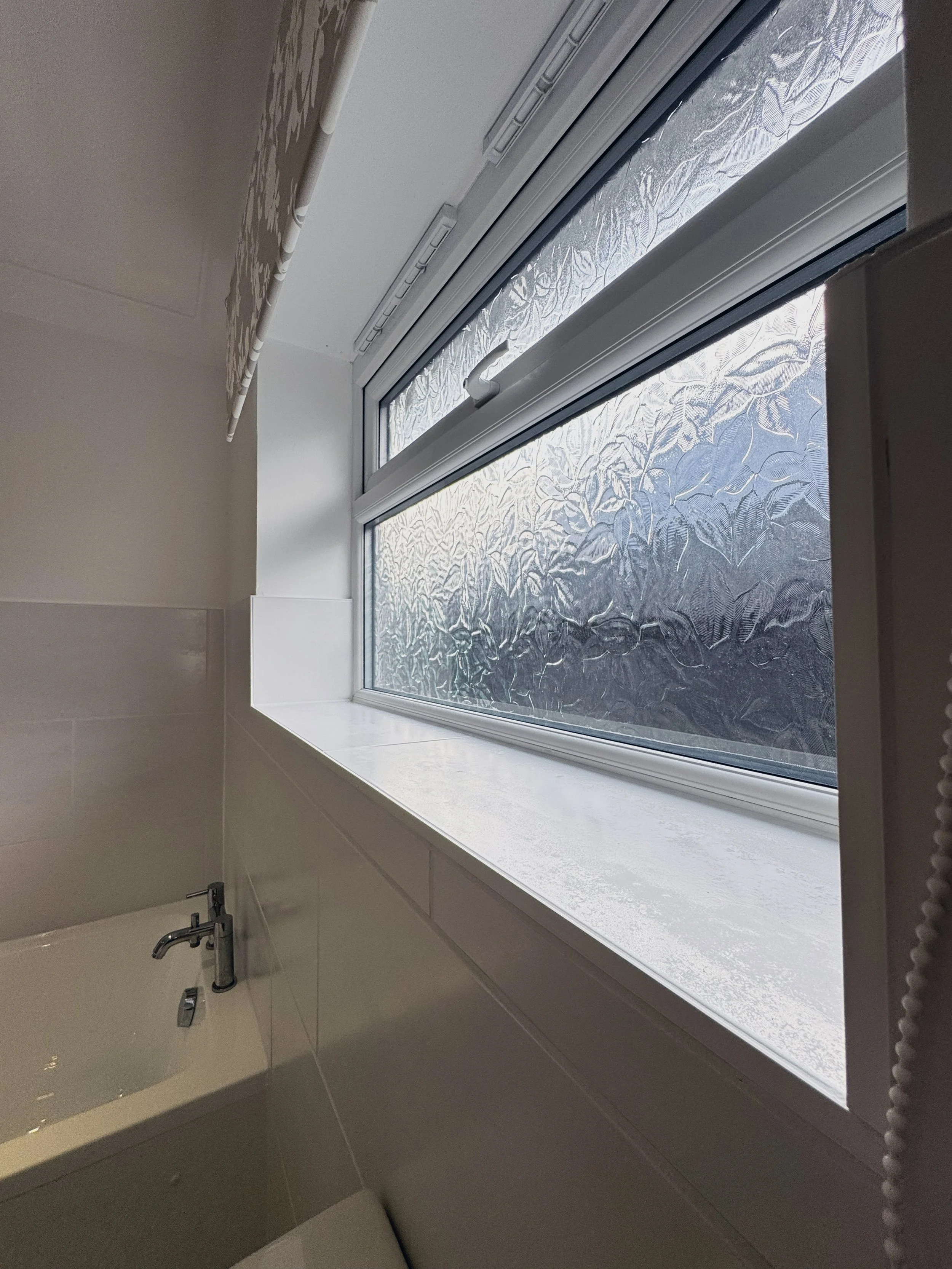 Frosted window above a bathtub in a bathroom, with a beige handle and beige tiled wall.