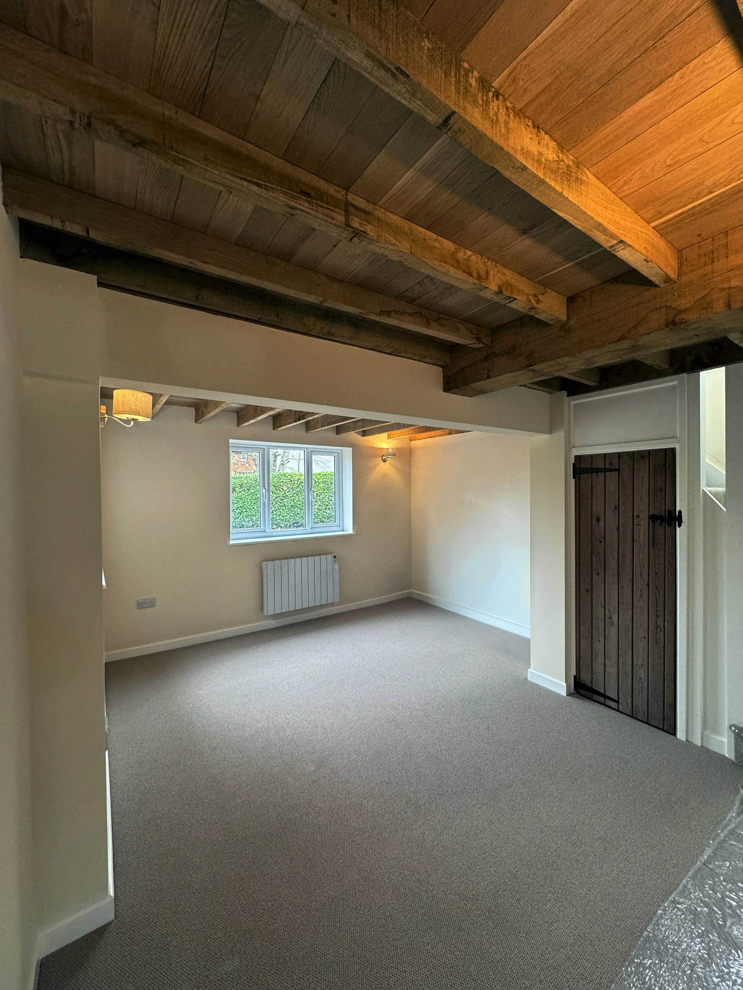 Empty room with wooden ceiling beams, a window with greenery outside, a radiator below the window, and a small, closed wooden door.