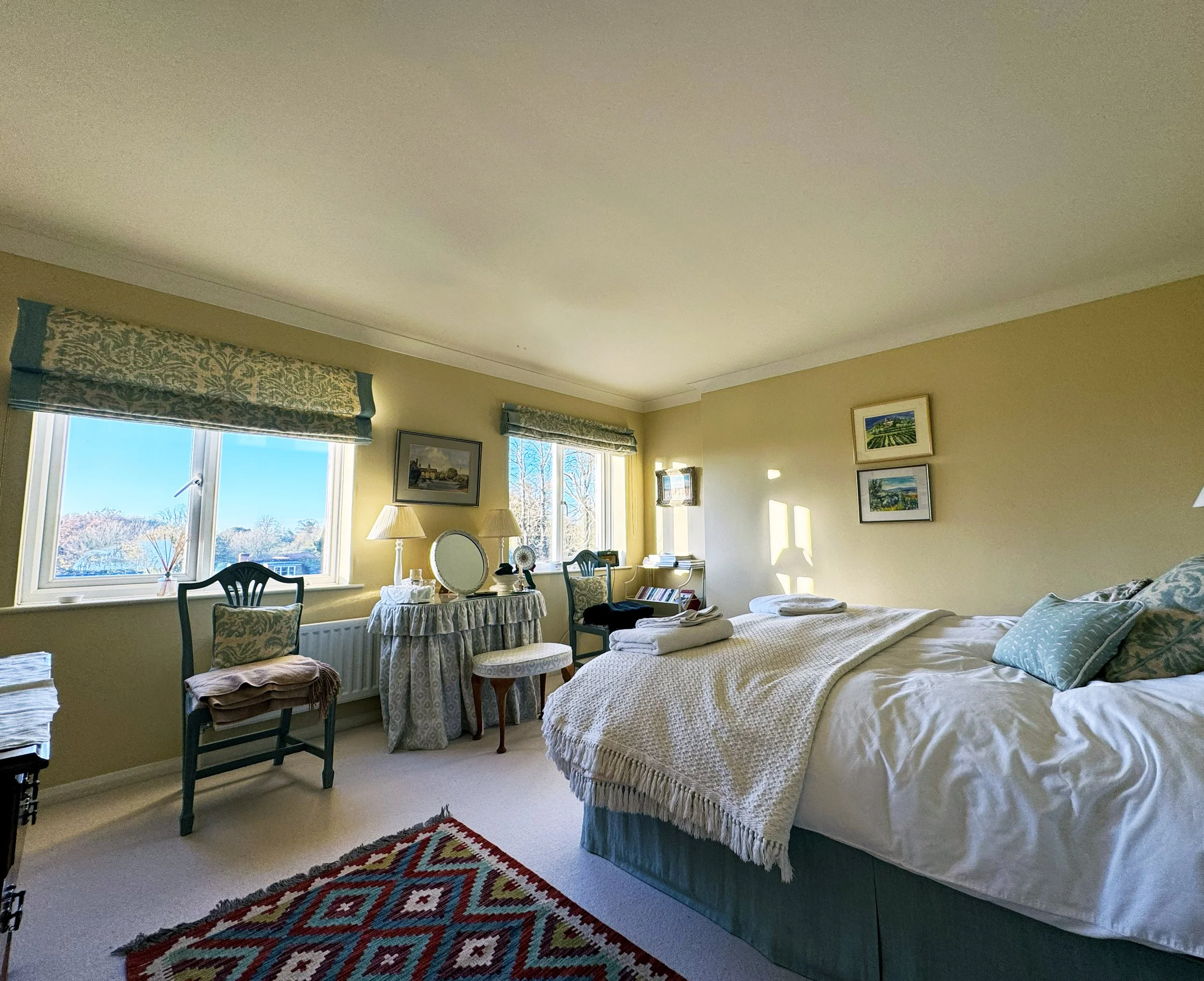 A cozy bedroom with a bed, a window view of trees and blue sky, and vintage furnishings including a round table with a mirror and lamps, and framed artwork on yellow walls.