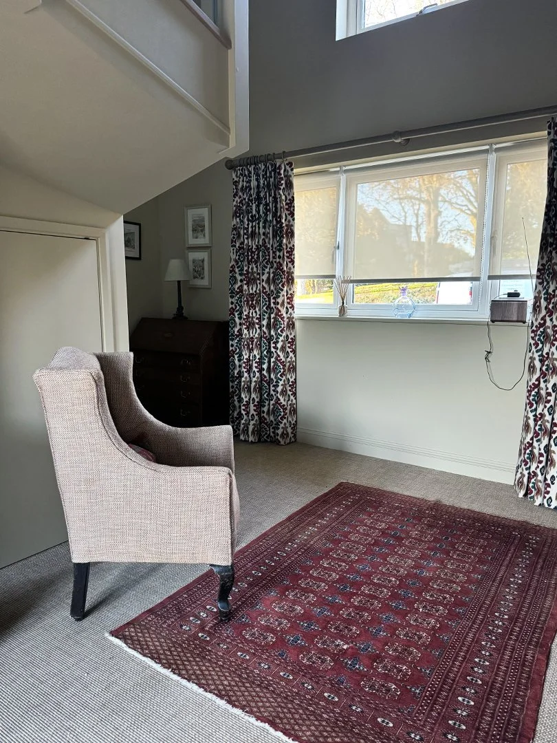 A cozy room with a beige armchair, a red patterned area rug, a window with floral curtains, and a wooden cabinet with a lamp, picture frames, and decorative items.